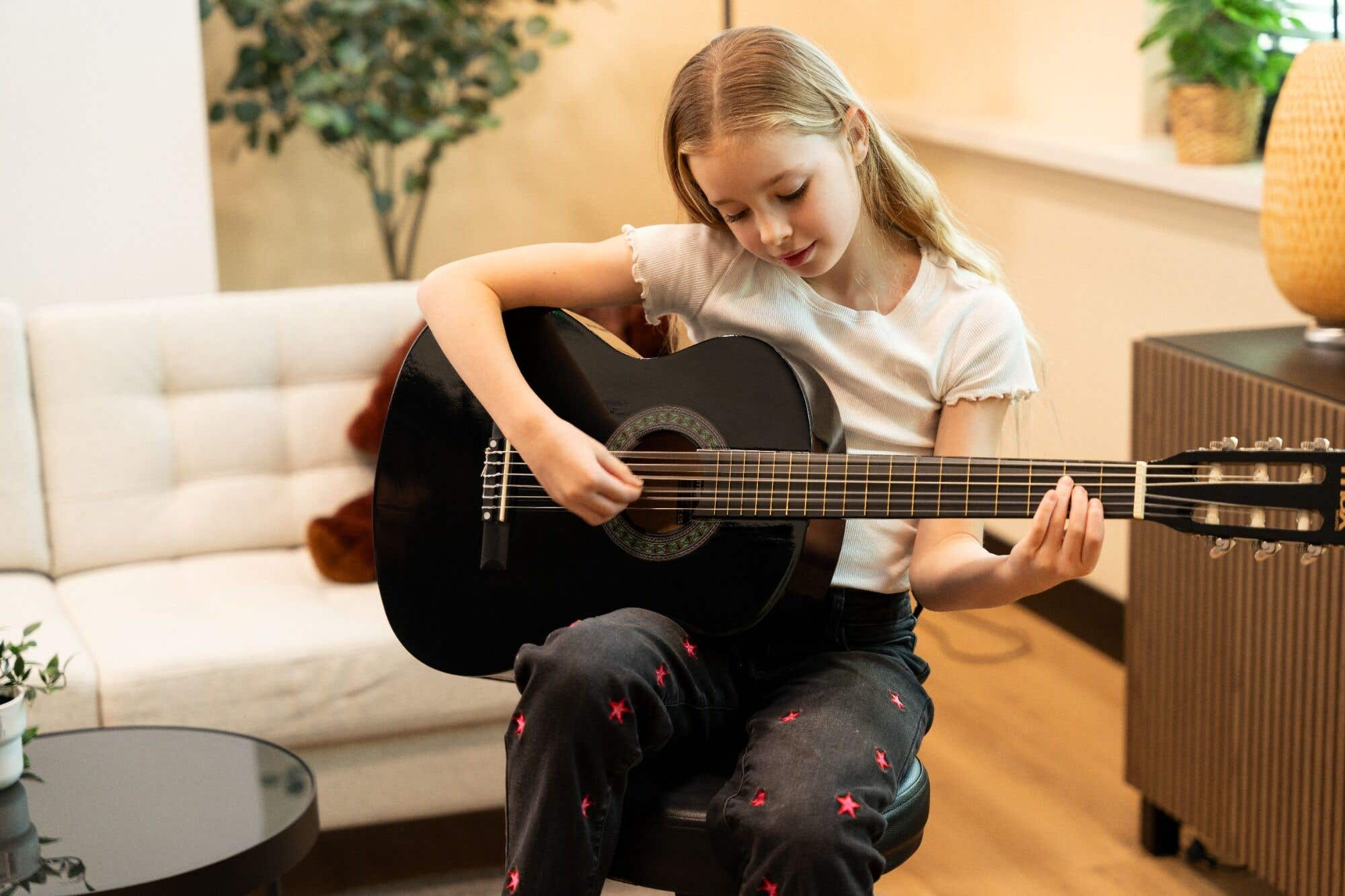 Een jong meisje met blond haar speelt gitaar op een zwarte gitaar terwijl ze op een kruk zit. Ze draagt een wit shirt en een spijkerbroek met sterren.