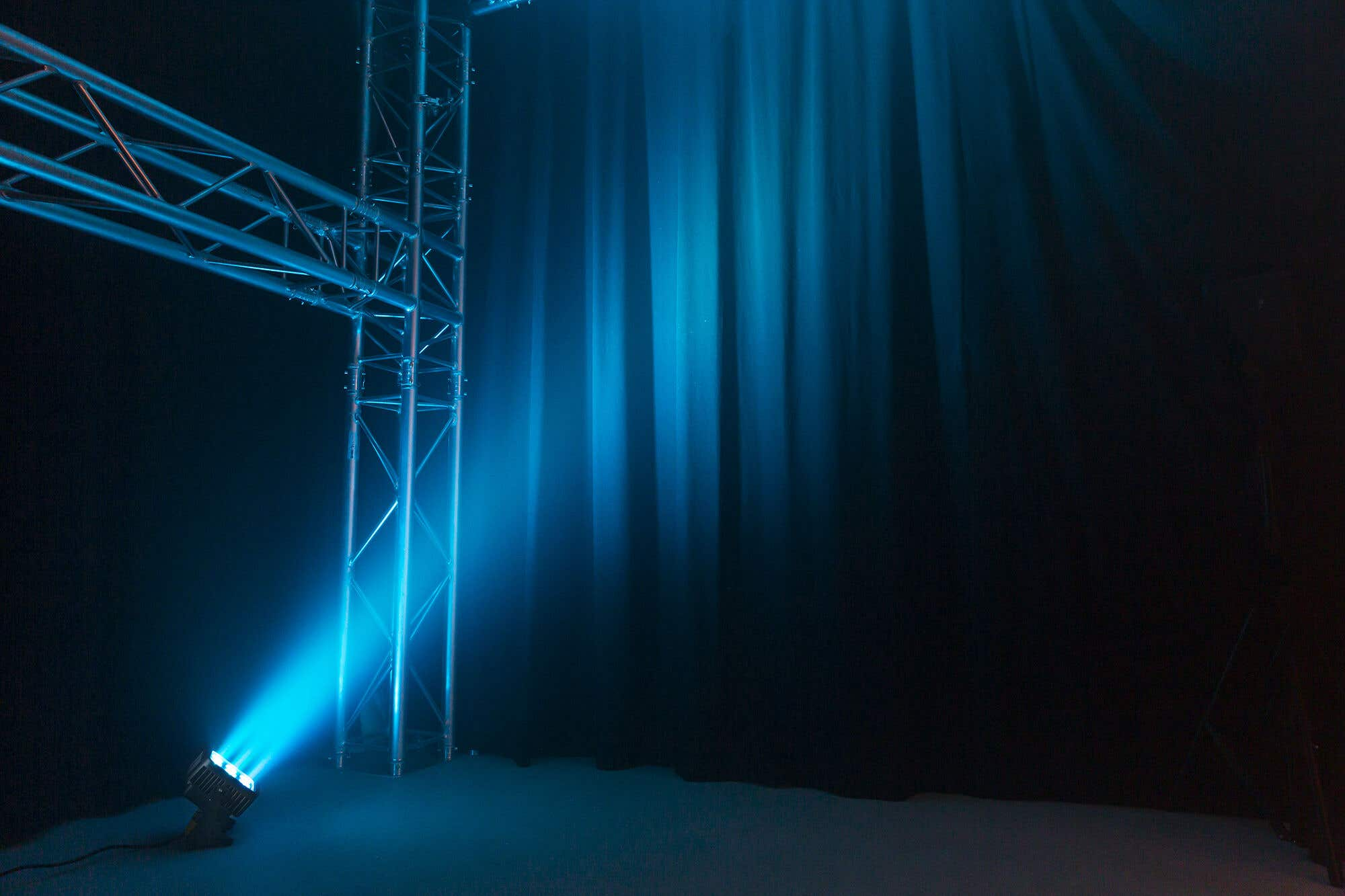 Un escenario con un foco azul que ilumina una estructura metálica y cortinas oscuras. El suelo es azul.