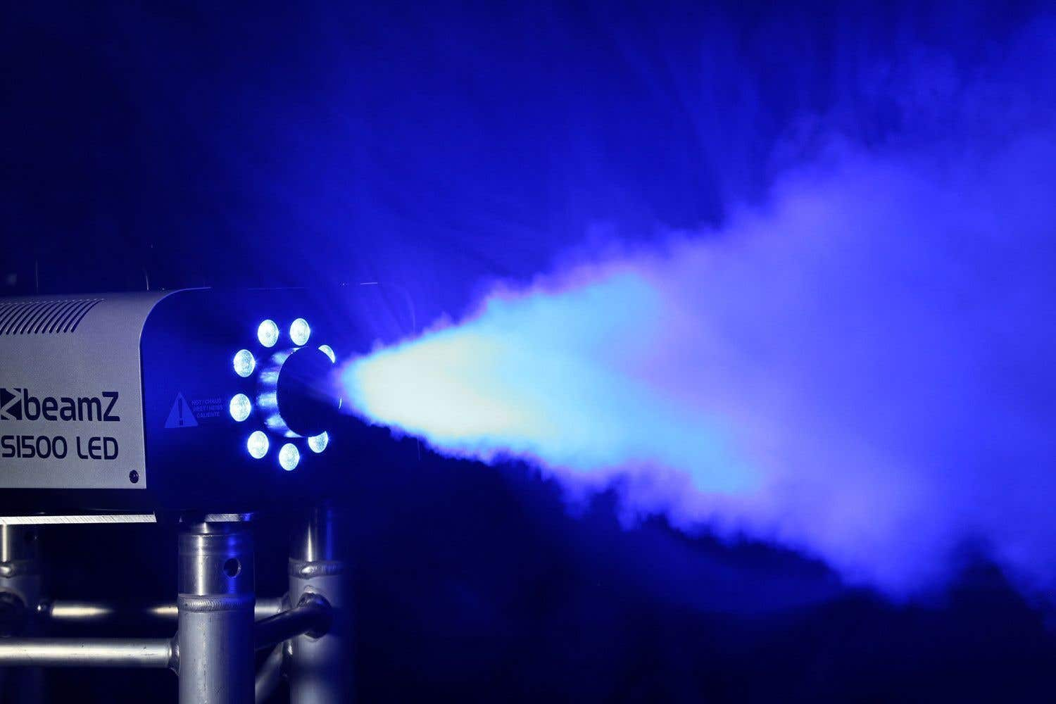 Una máquina de niebla beamZ S1500 LED emite humo azulado. Sus luces brillan, el texto es visible sobre un fondo oscuro.