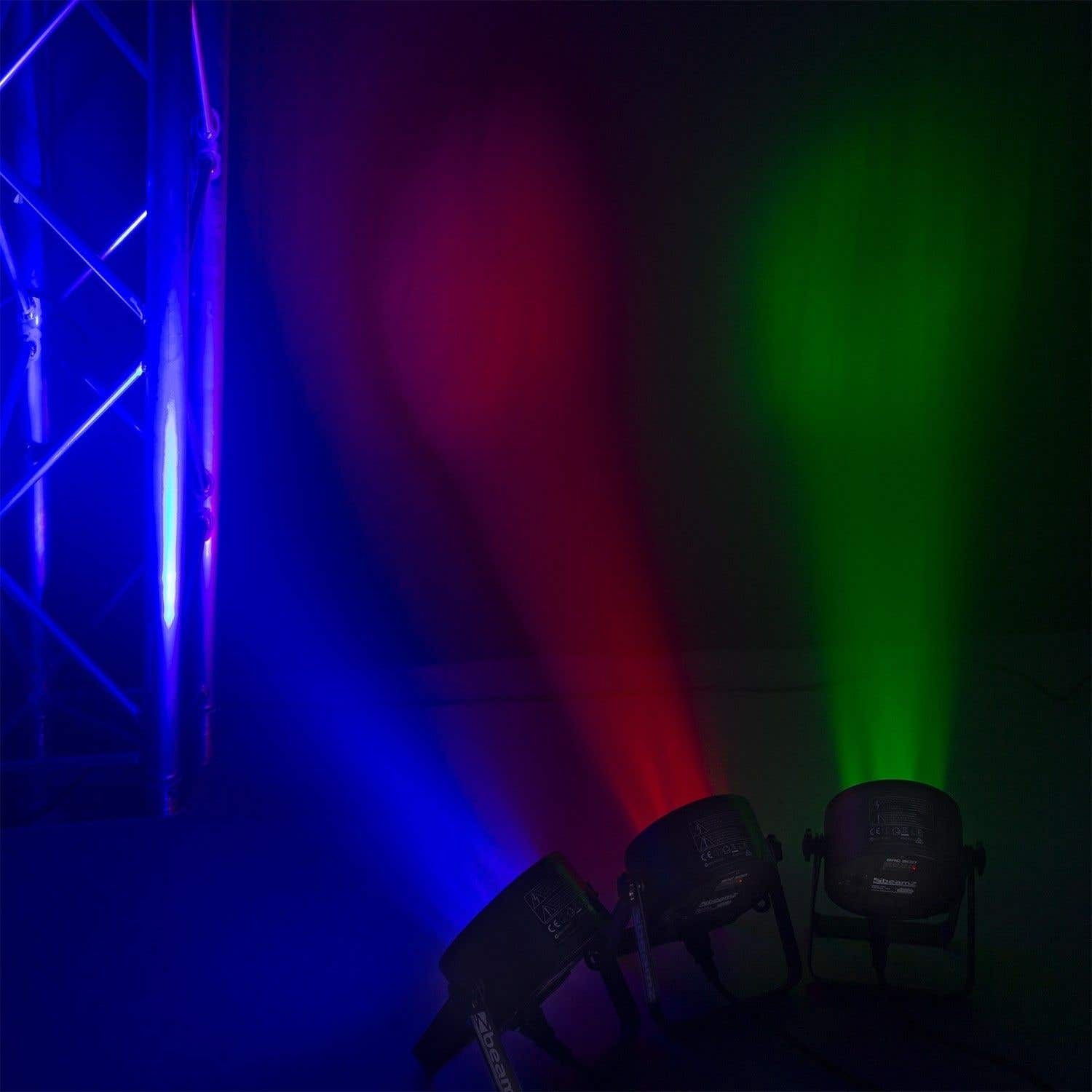 Tres luces de escenario proyectan haces azules, rojos y verdes. Una estructura metálica está a la izquierda. El fondo es oscuro.