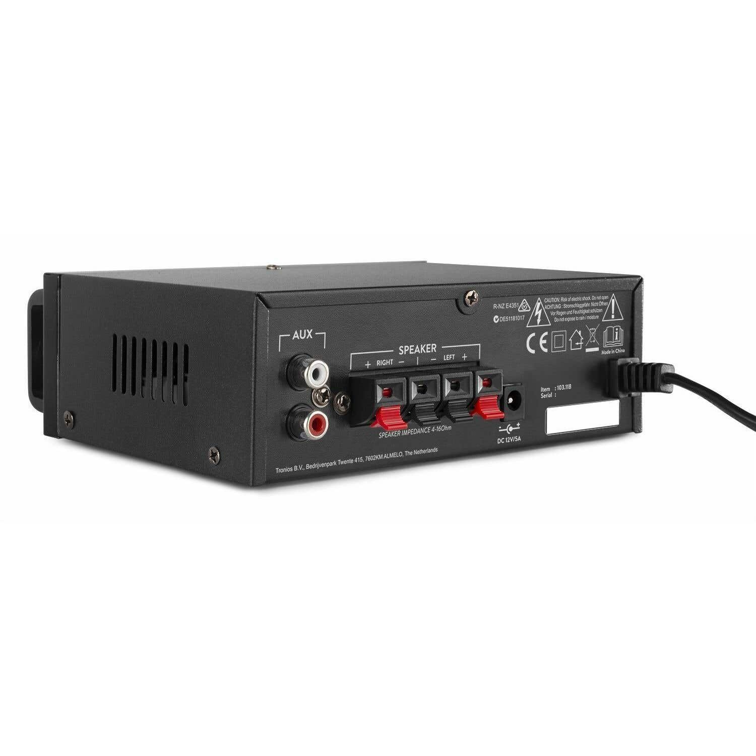 Amplificador de audio negro con entradas, terminales de altavoz y etiquetas de advertencia. Cable de alimentación enchufado. Texto incluye 'AUX', 'SPEAKER', y 'DC 12V/5A'.