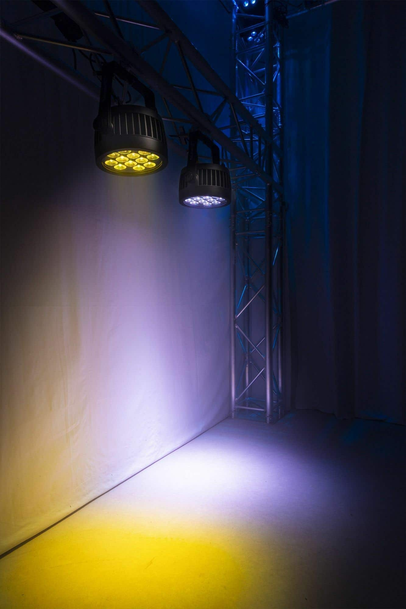 Configuración de iluminación escénica con dos focos que proyectan luz amarilla y púrpura. Se ve una estructura metálica y un fondo blanco.