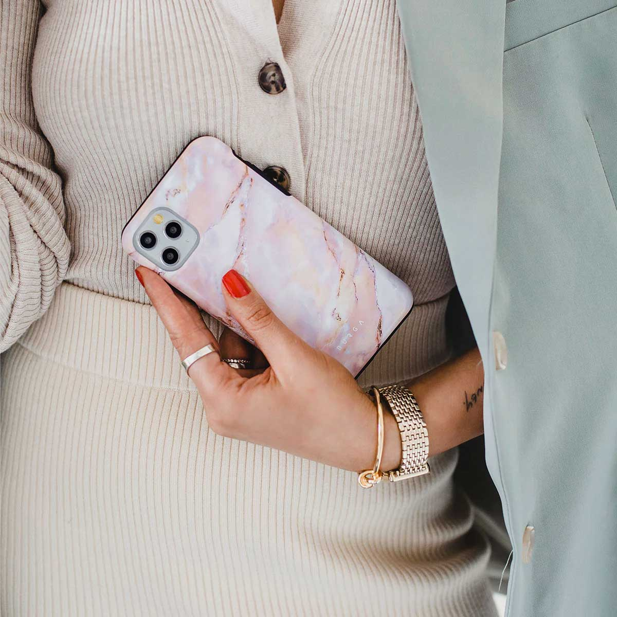 Vrouw die een telefoon vasthoudt met een marmeren hoesje. Ze draagt een lichte trui, ringen, een armband en een lichtgroene jas.