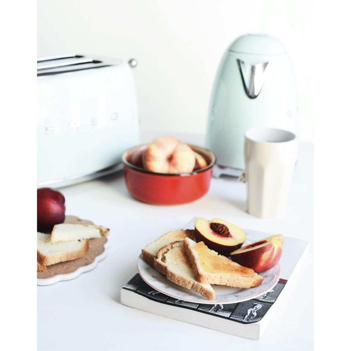 Scena śniadaniowa: Toster, czajnik, kubek, tosty, brzoskwinie i czerwona miska na białym stole. Chleb na dekoracyjnym talerzu.