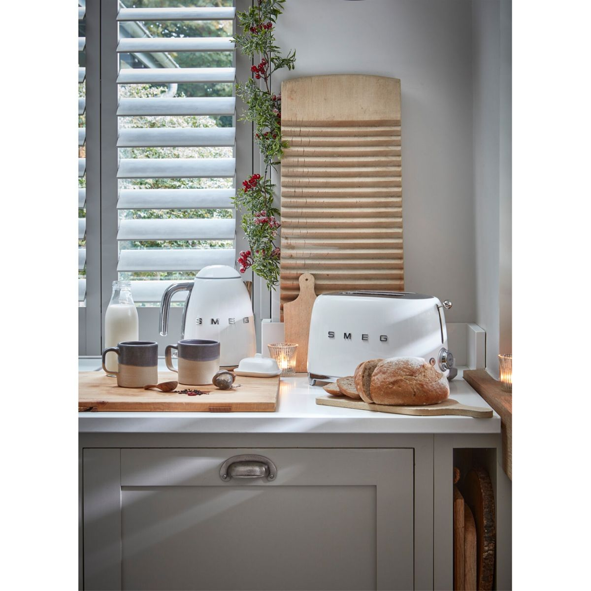 Scena kuchenna: Biały czajnik i toster, kubki, chleb i drewniane deski do krojenia na blacie. Żaluzje w tle.
