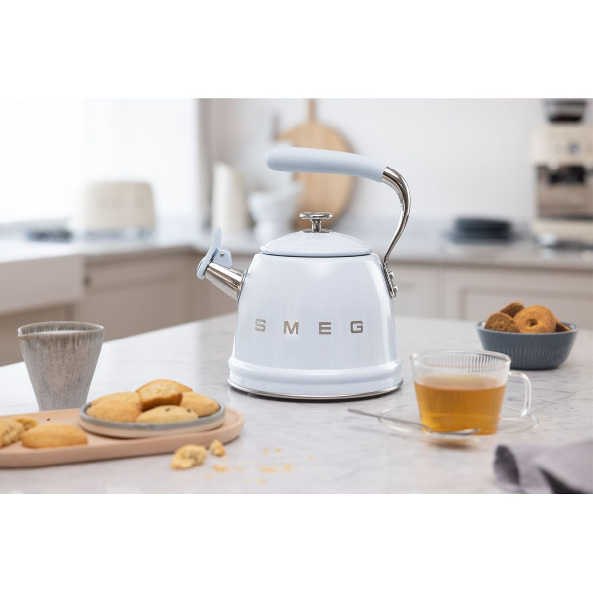 Ein hellblauer SMEG-Wasserkocher, Tee, Kekse und eine Tasse stehen auf einer Marmortheke. Eine Schüssel mit Keksen und eine graue Tasse sind ebenfalls vorhanden.