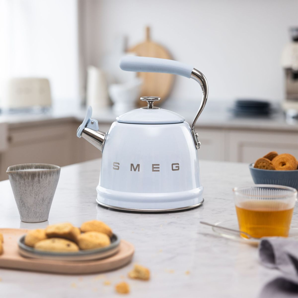 Ein hellblauer SMEG-Wasserkocher, Kekse, eine Tasse Tee und eine kleine Tasse stehen auf einem Marmortisch. Im Hintergrund ist eine Küche zu sehen.