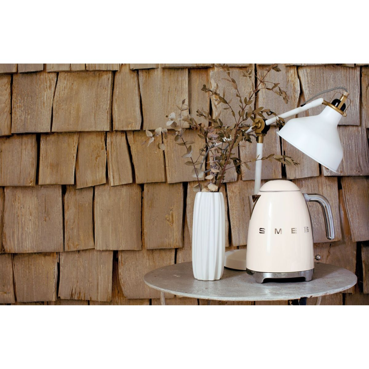 Kremowy czajnik Smeg, biała lampa i biały wazon z gałązkami na małym stoliku, drewniane tło.
