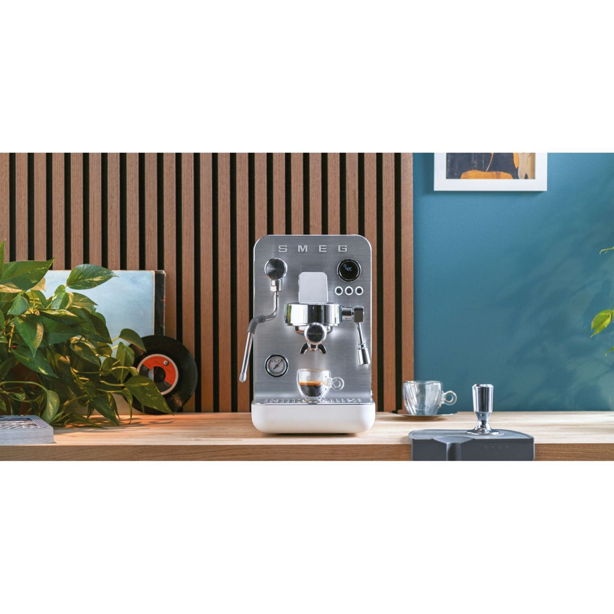 Eine Edelstahl-Espressomaschine von SMEG mit einem Espresso. Ein Holztisch mit Pflanzen und einer Schallplatte im Hintergrund.