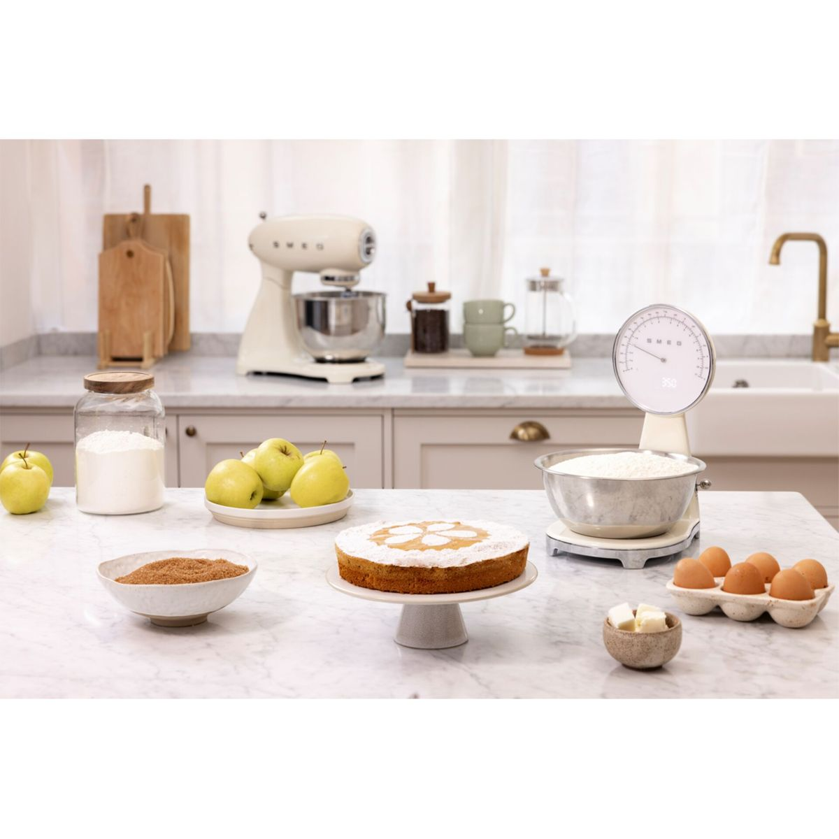 Scena kuchenna ze składnikami do pieczenia. Ciasto, jabłka, mąka, jajka i mikser na marmurowym blacie. Obecna jest również waga.