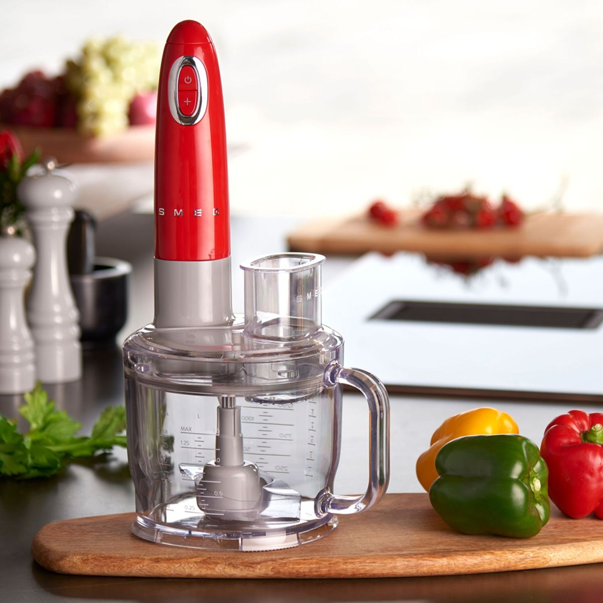 Czerwony i szary robot kuchenny na drewnianej desce z papryką. W tle inne składniki i przedmioty kuchenne.