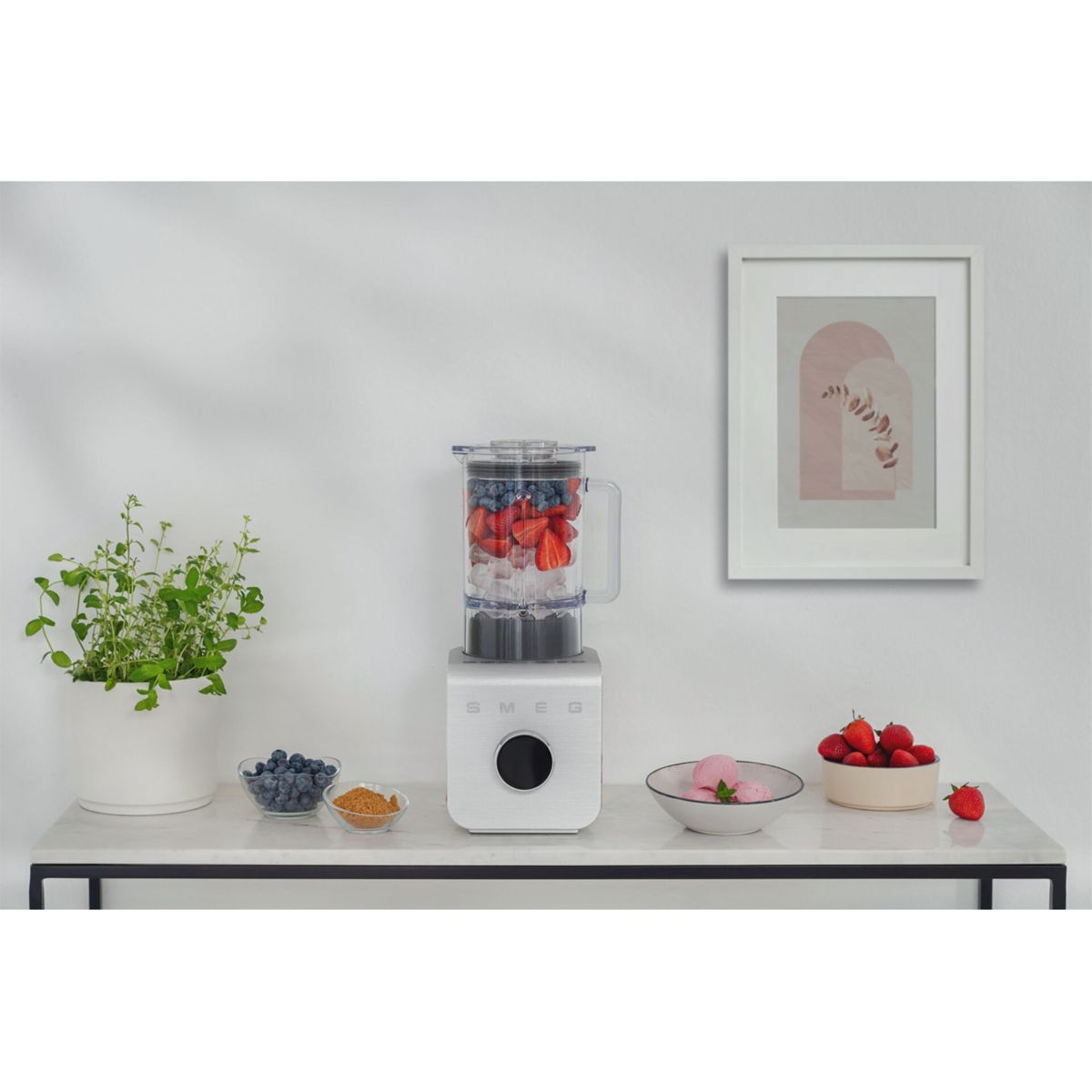 Un frullatore bianco con fragole e mirtilli è su un bancone. Altre ciotole contengono bacche e gelato. Un quadro incorniciato è appeso al muro.