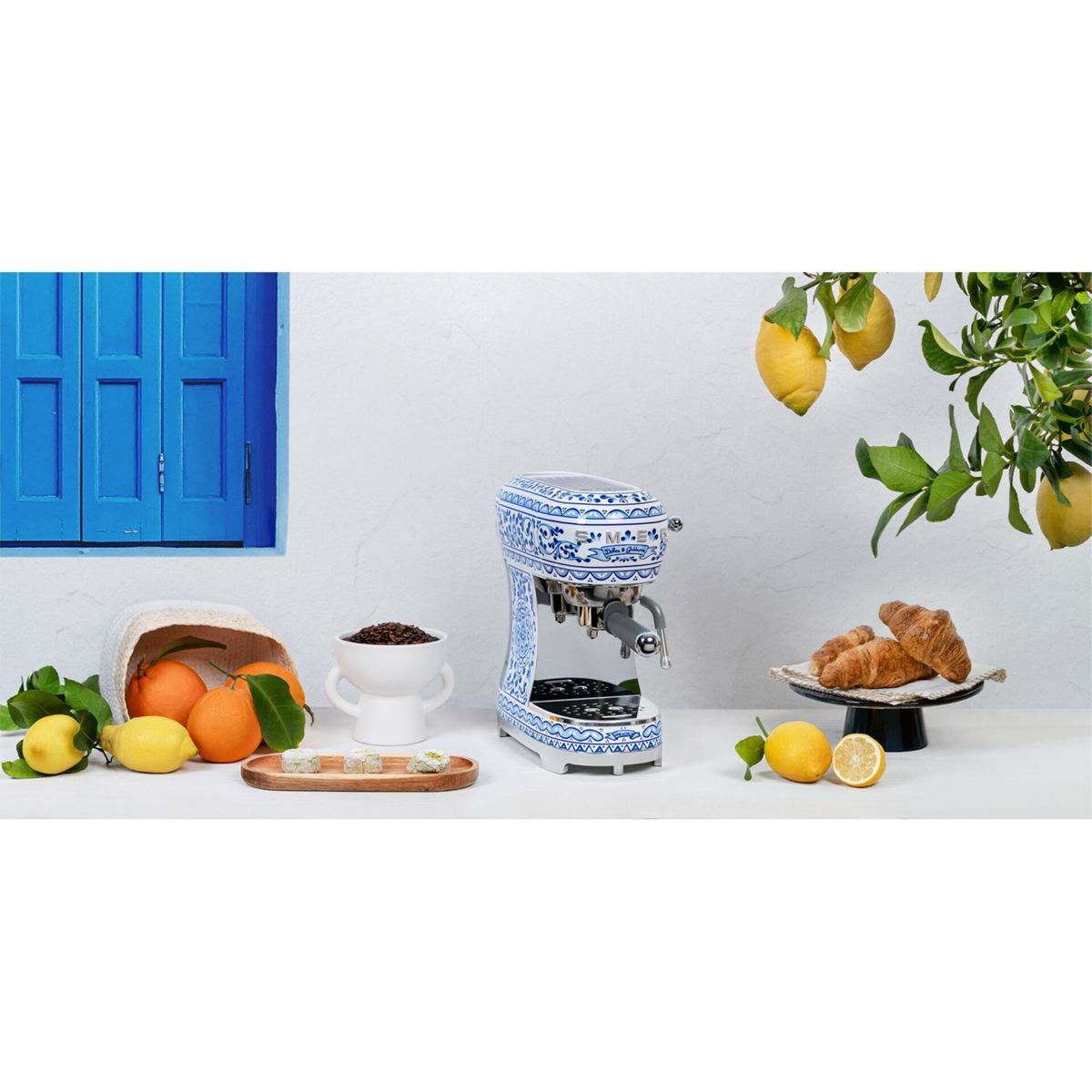Eine blau-weiße Espressomaschine mit Orangen, Zitronen, Croissants und Kaffeebohnen. Im Hintergrund befindet sich ein blauer Fensterladen.