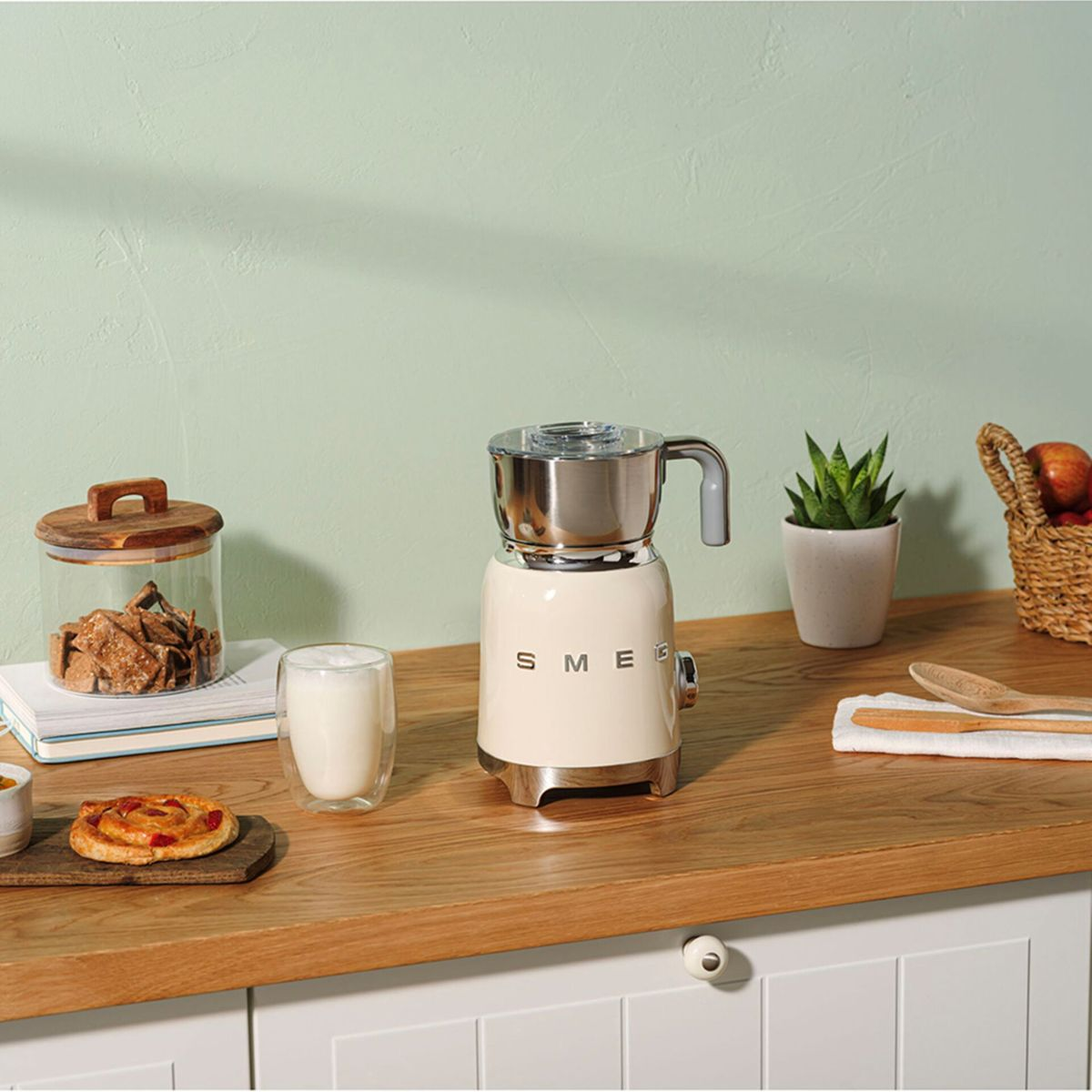 Scena na blacie kuchennym z spieniaczem do mleka SMEG, szklanką mleka, ciastkami, ciasteczkami i rośliną. Drewniana powierzchnia i biała szafka.