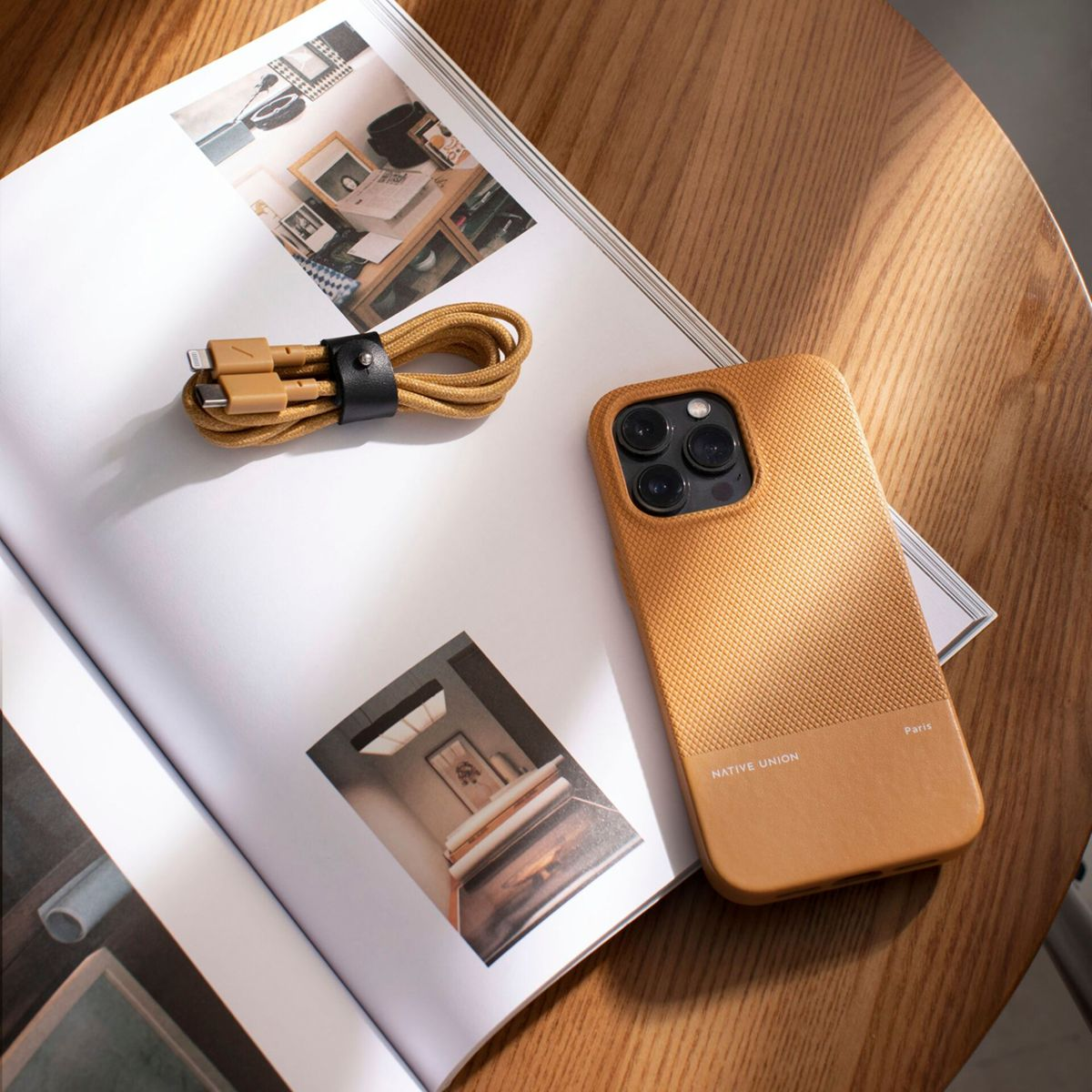 Ein Handycase, ein Kabel und eine Zeitschrift liegen auf einem Holztisch. Das Handycase ist braun und das Kabel goldfarben. Eine Zeitschrift ist geöffnet.