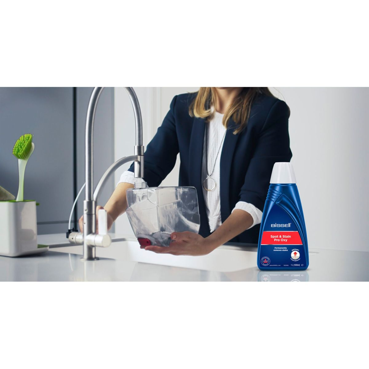 Een vrouw vult een doorzichtige container in een gootsteen. Een fles Bissell Spot & Stain Pro Oxy staat op het aanrecht.
