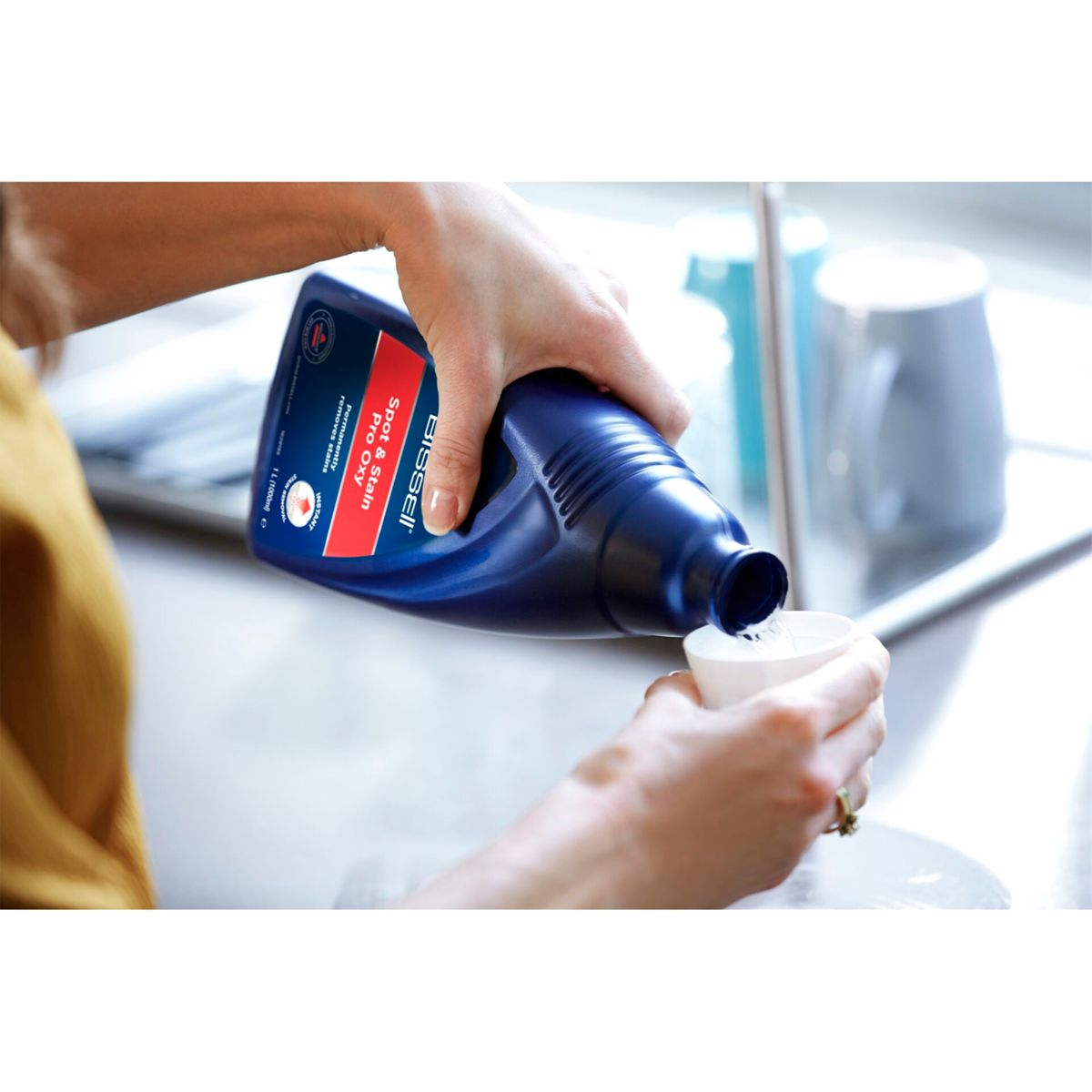 Een persoon giet blauwe vloeistof uit een Bissell-fles in een witte maatbeker. De scène is een spoelbak in de keuken.