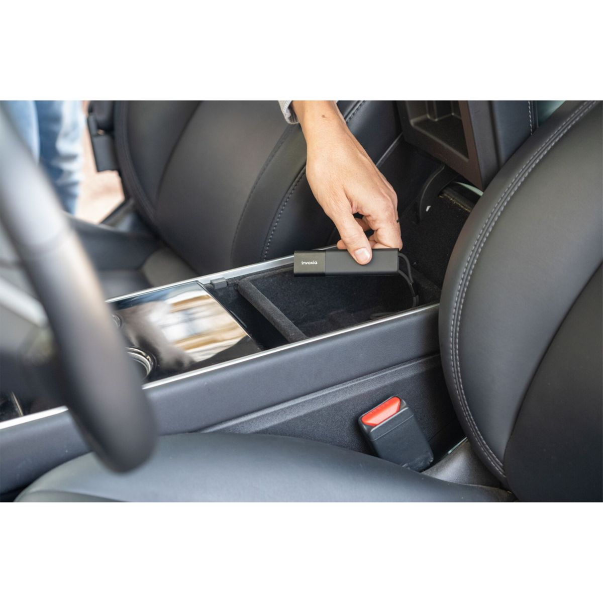 Ręka umieszczająca czarne urządzenie z napisem 'Invoxia' w konsoli środkowej samochodu. Widoczne czarne skórzane siedzenia i klamra pasa bezpieczeństwa.