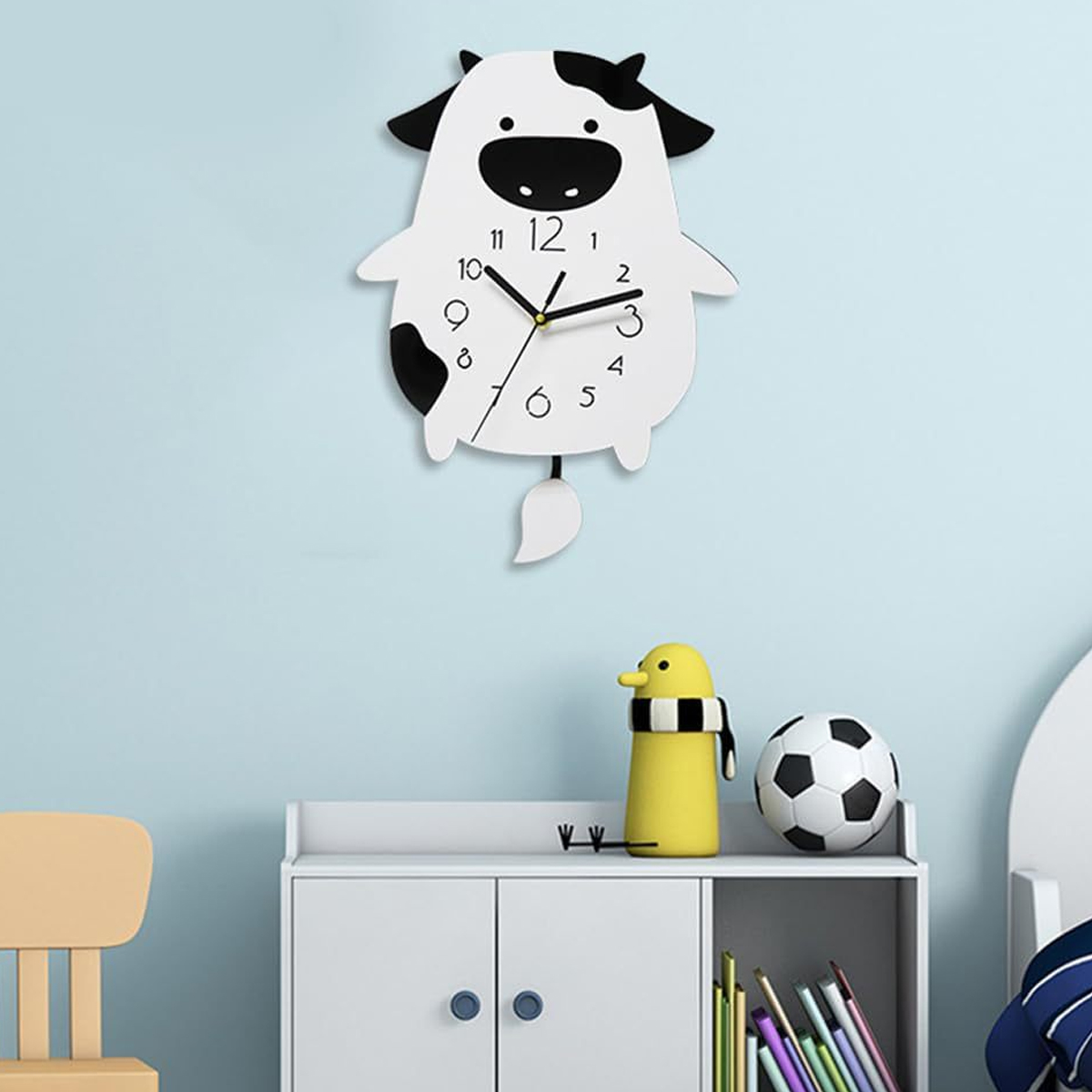 Eine kuhförmige Uhr hängt an einer blauen Wand. Darunter befinden sich ein gelber Pinguin, ein Fußball und Bücher.
