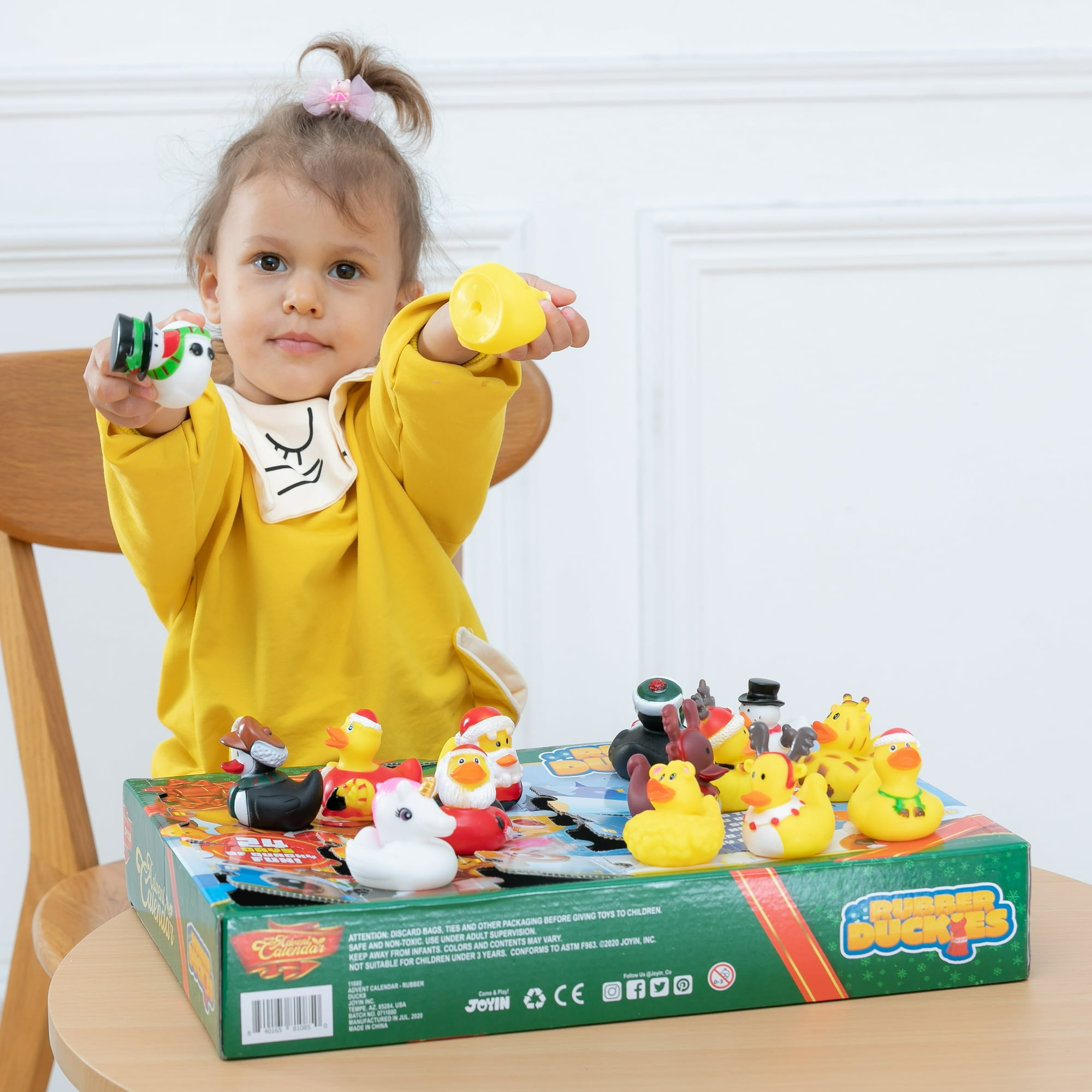 Ein Kind hält Gummienten-Spielzeug und eine Schneemann-Ente. Auf dem Tisch steht eine Box mit Gummienten im Weihnachtsthema.