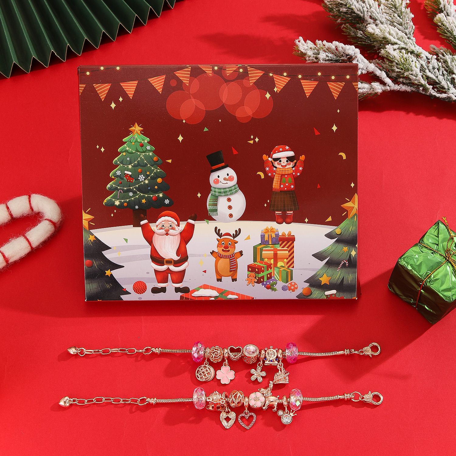 Weihnachtsschachtel mit Weihnachtsmann, Schneemann und Ornamenten. Zwei Charm-Armbänder liegen auf rotem Untergrund.