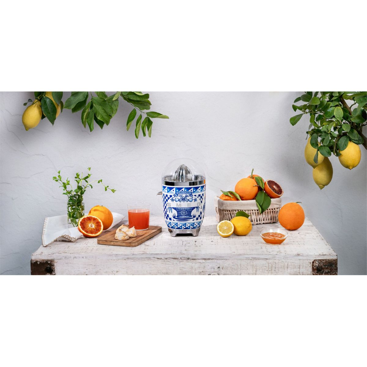Ein Tisch mit Orangen, Zitronen und einer Saftpresse. Ein Glas Saft und ein Korb mit Orangen sind ebenfalls vorhanden, zusammen mit Laub.