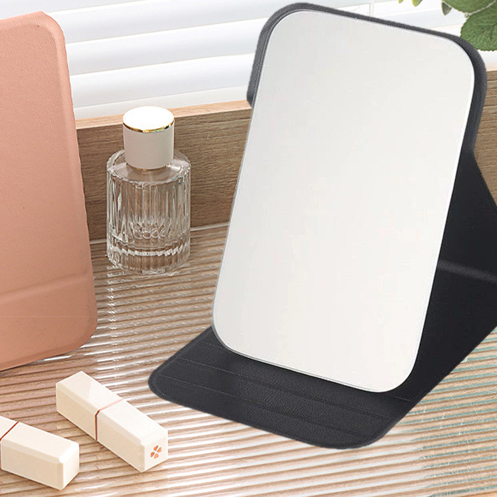 Ein Kompaktspiegel ist auf einem Tisch geöffnet. Eine Parfümflasche und zwei Lippenstifte sind in der Nähe. Der Hintergrund umfasst ein rosa Etui.