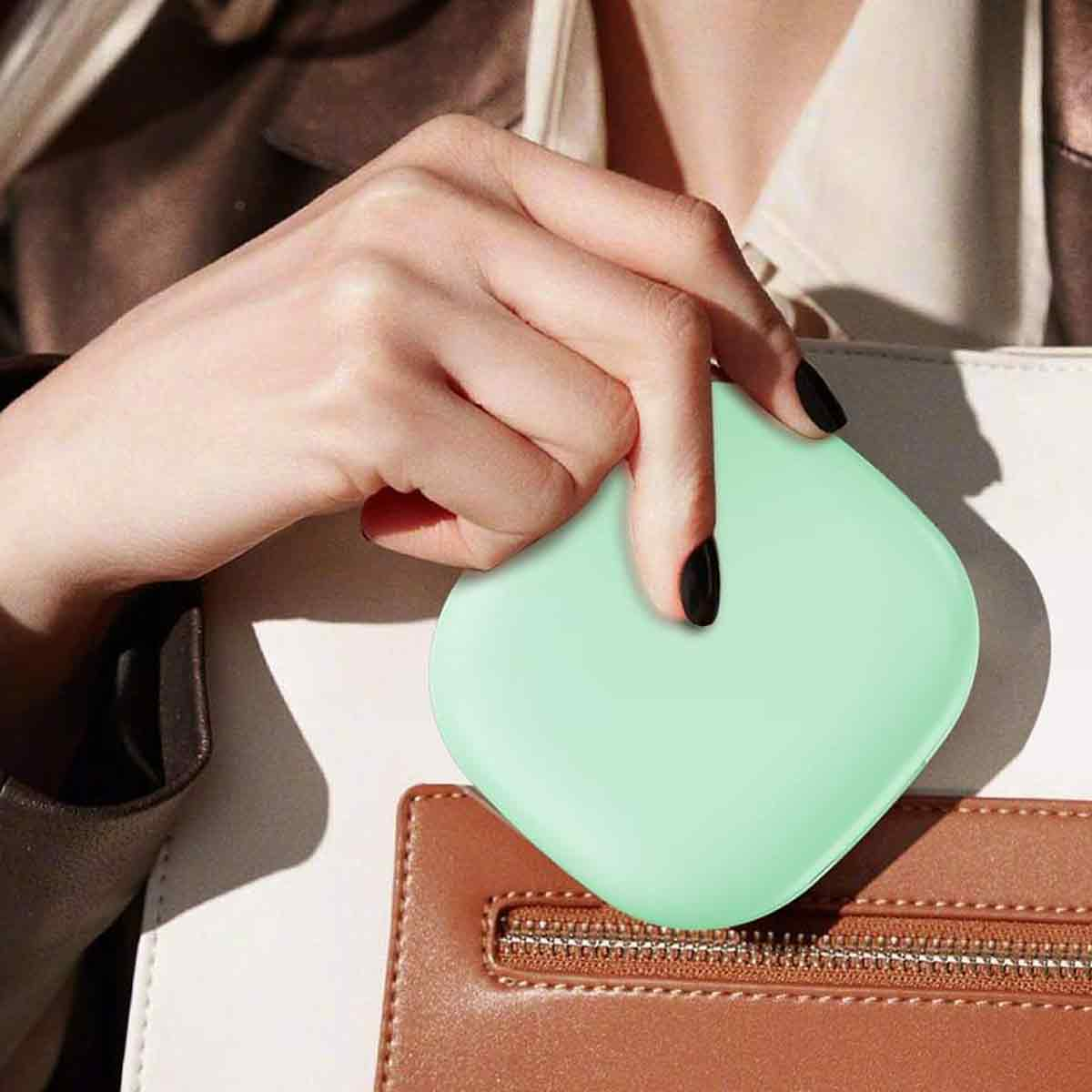Eine Hand hält ein mintgrünes, quadratisches Objekt, möglicherweise einen Kompaktspiegel, über einer braunen Handtasche. Die Nägel sind schwarz lackiert.