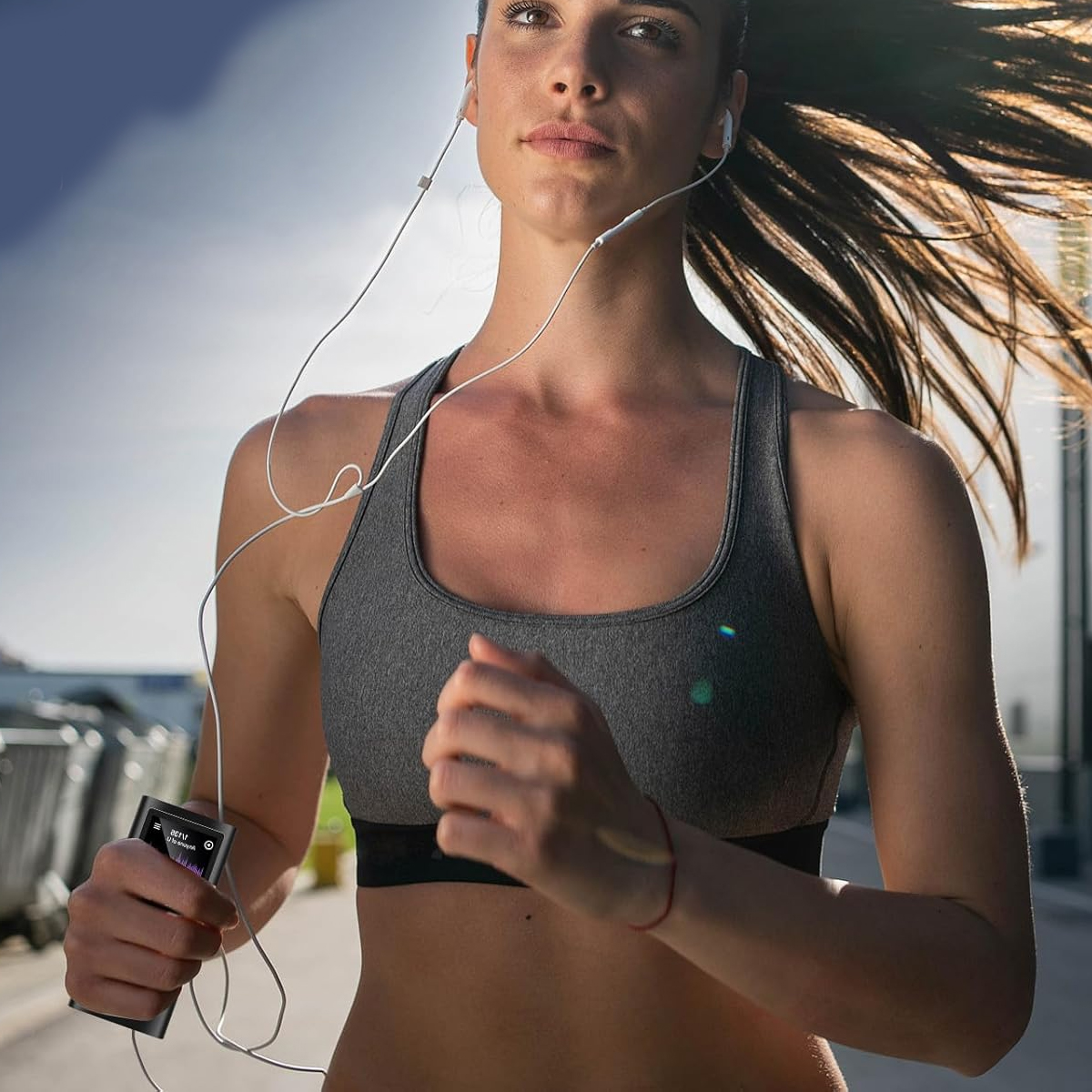 Eine Frau läuft draußen, trägt einen grauen Sport-BH und Kopfhörer. Sie hält einen Musik-Player. Ihr Haar weht. Der Hintergrund ist unscharf.