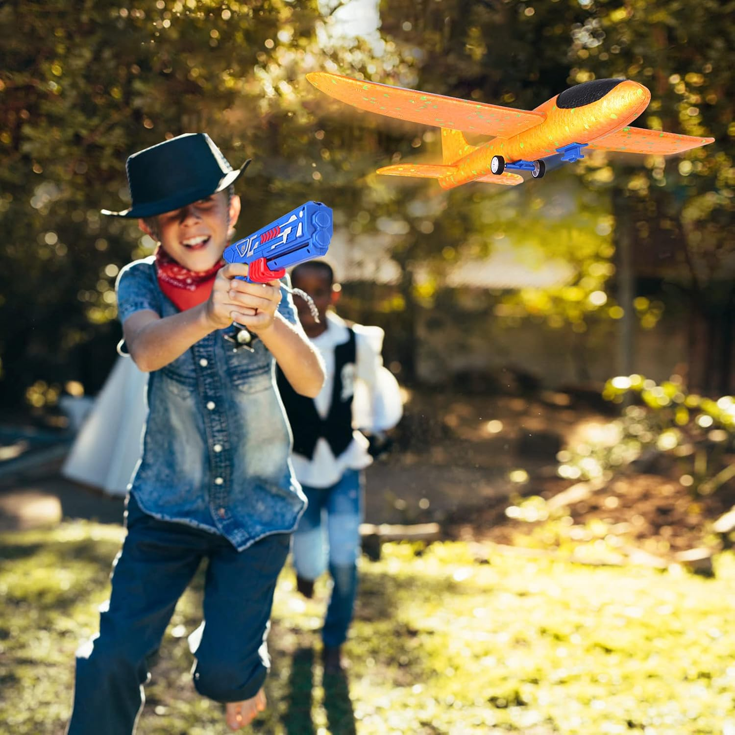 Ein Junge mit Spielzeugpistole und Hut zielt auf ein Spielzeugflugzeug. Ein anderer Junge rennt im Hintergrund. Szene ist draußen an einem sonnigen Tag.