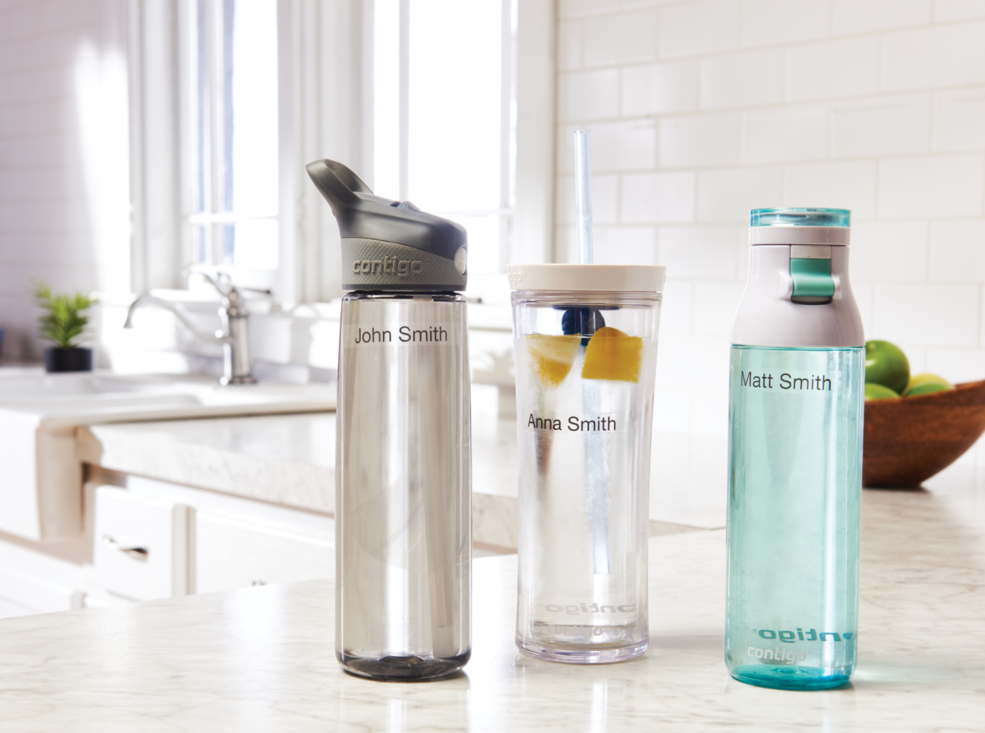 Trzy butelki z wodą z imionami 'John Smith', 'Anna Smith' i 'Matt Smith' na blacie kuchennym z miską jabłek.