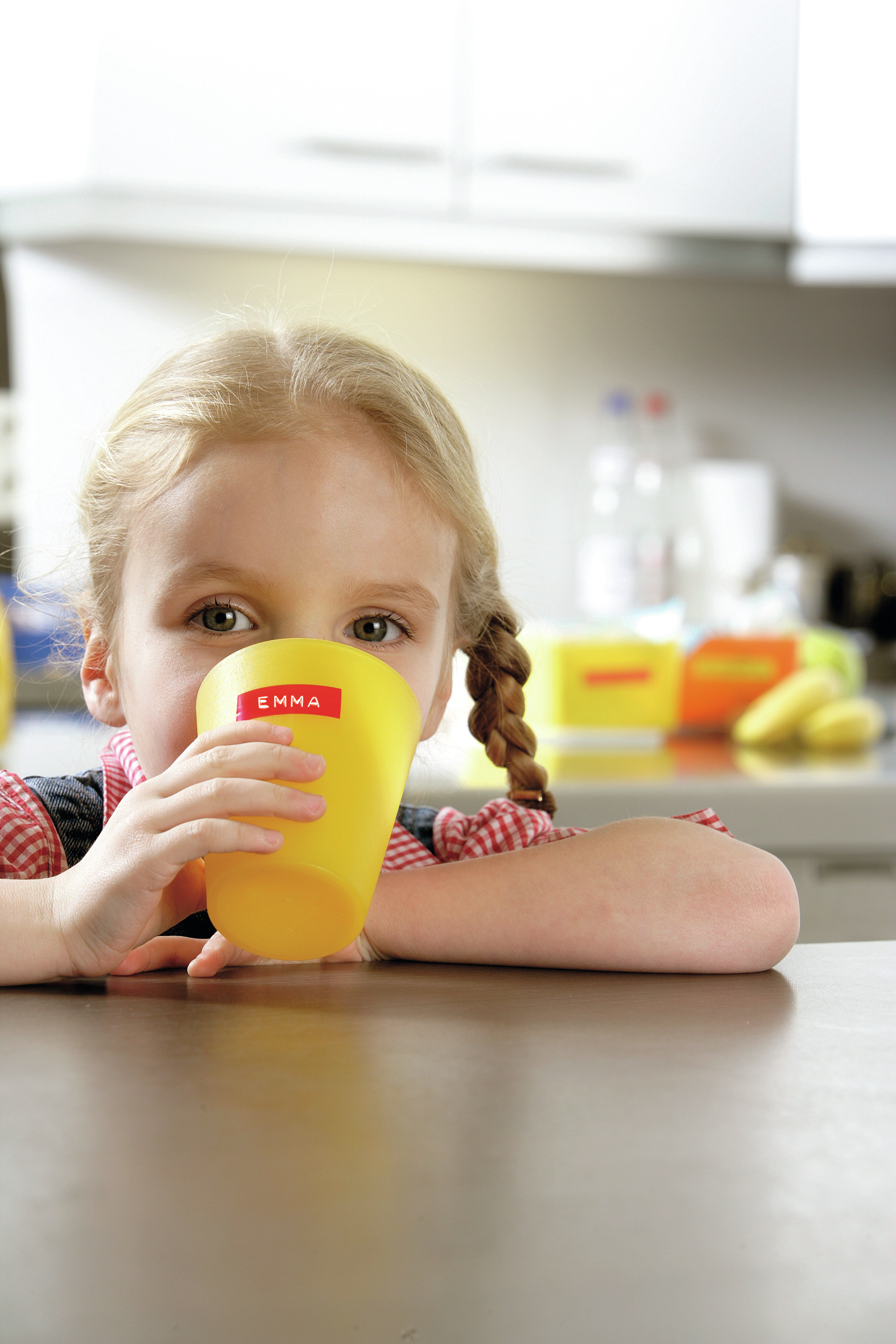Dziewczynka pije z żółtego kubka z napisem "EMMA". Ma blond warkocze i nosi czerwoną i białą koszulę w kratę.