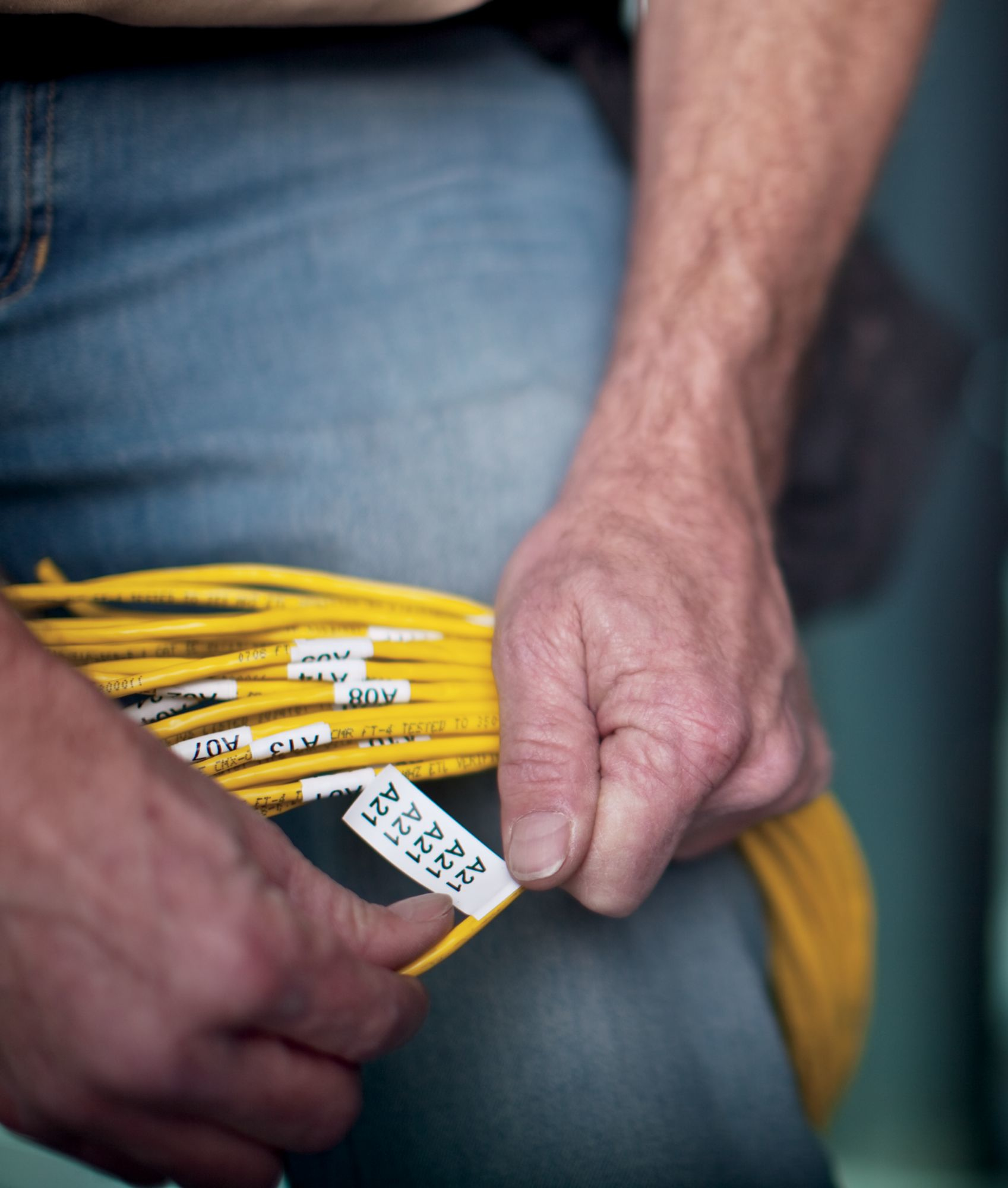 A person holds yellow cables with white labels. The person is wearing blue jeans. The labels have alphanumeric codes.
