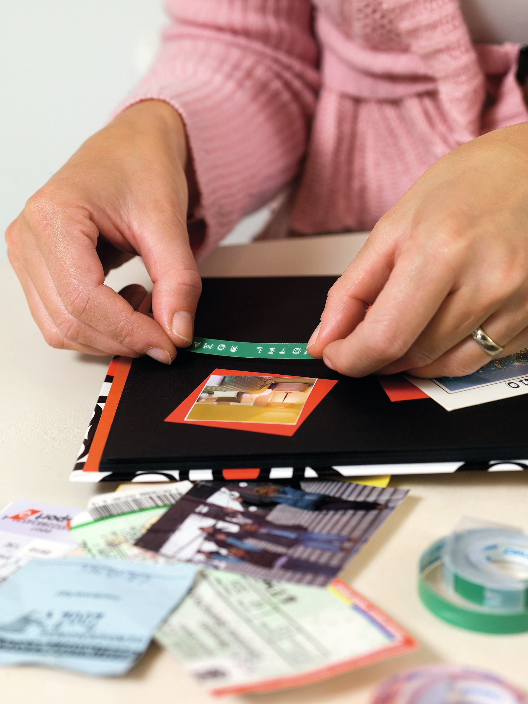 Ręce umieszczające zieloną naklejkę z napisem 'HOTEL ROMA' na albumie, ze zdjęciami, biletami i taśmą.