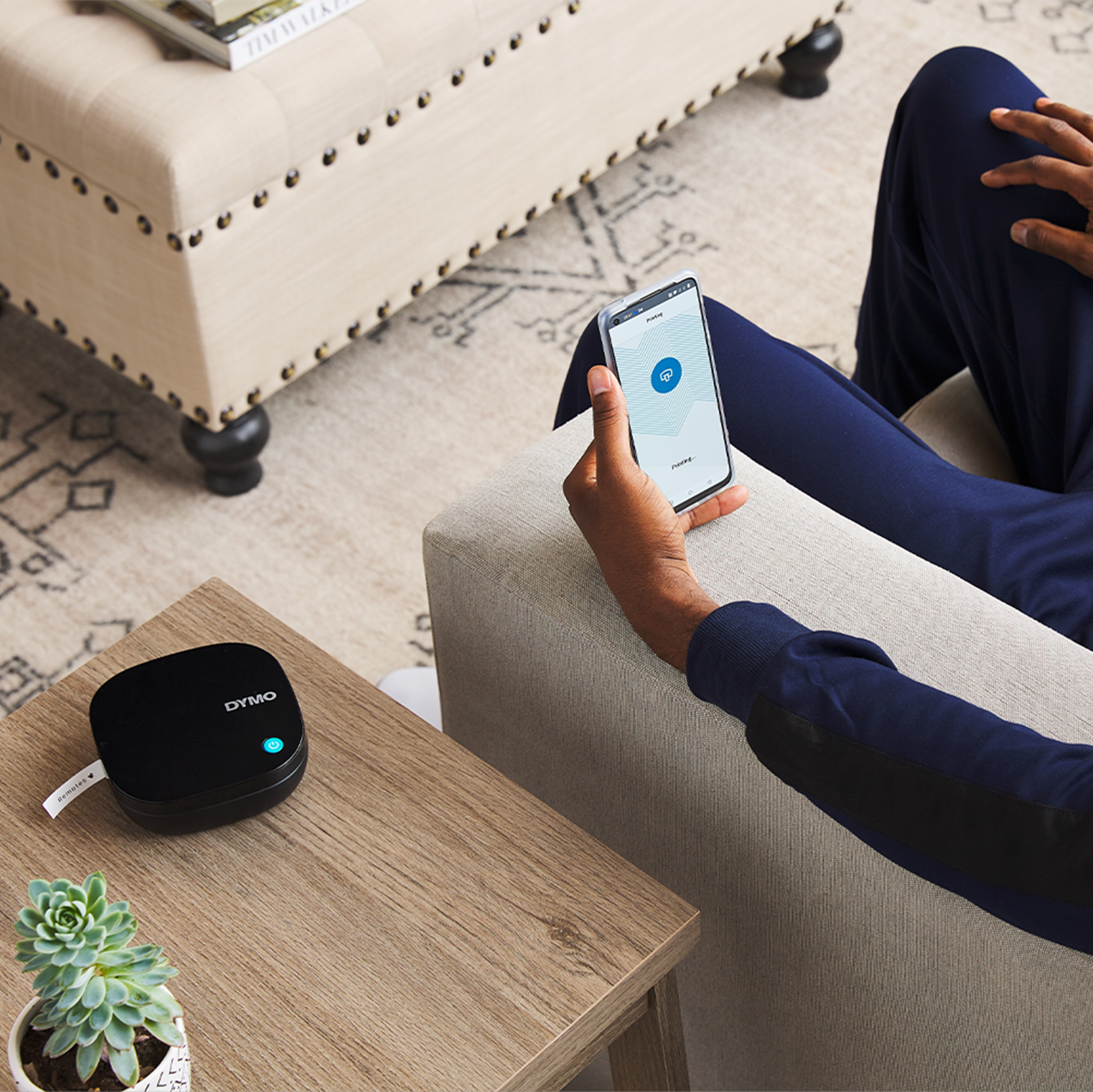 Osoba siedzi, używa telefonu. Czarna drukarka etykiet na stole. Sukkulent w białej doniczce na stole.