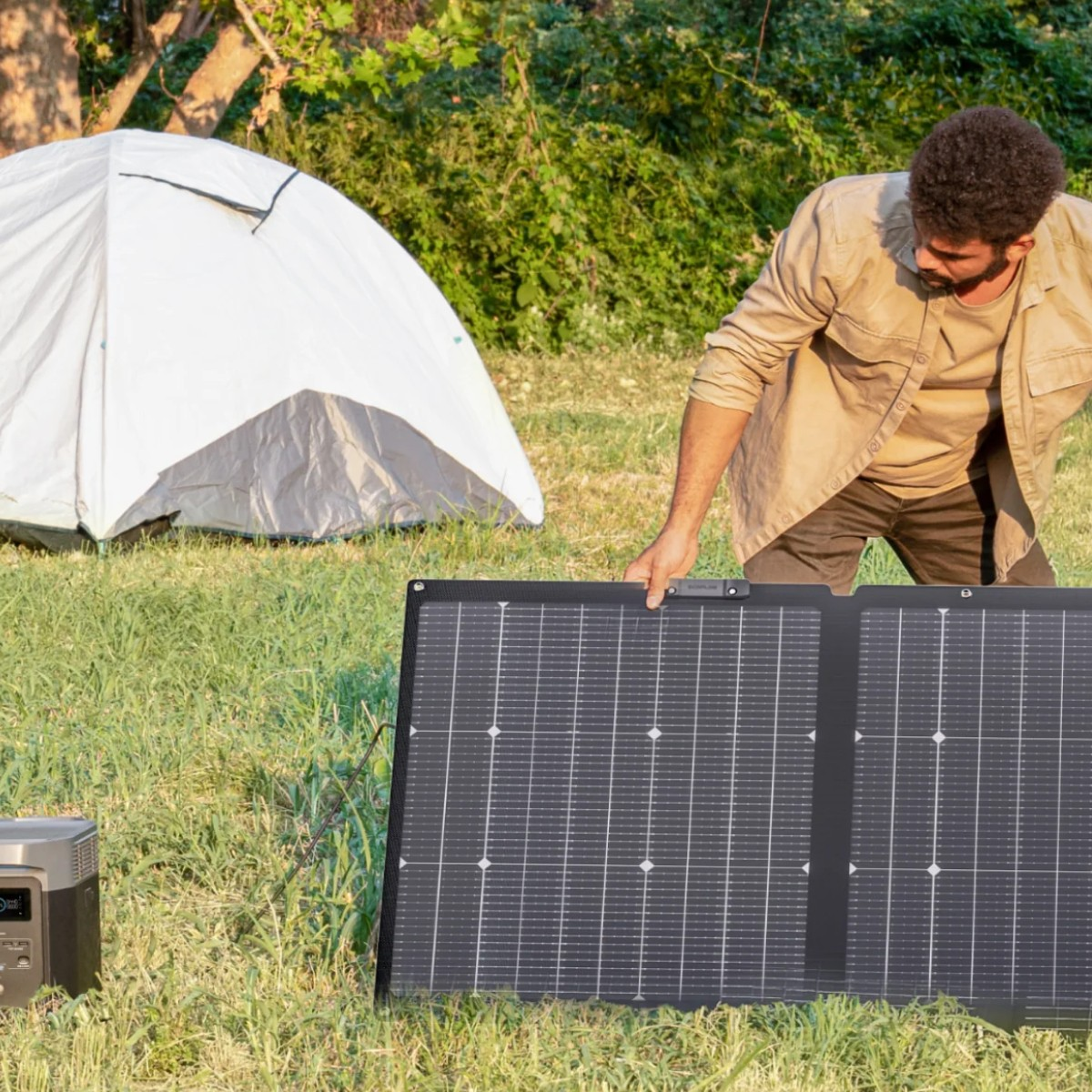Un hombre instala un panel solar cerca de una tienda de campaña en un campo de hierba. Una estación de energía está a la izquierda. Árboles en el fondo.