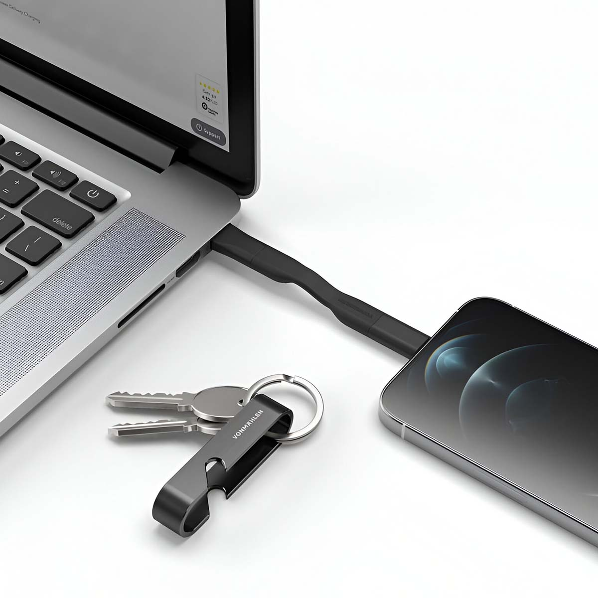 Laptop collegato a un telefono tramite un cavo nero. Chiavi e portachiavi apribottiglie in primo piano su superficie bianca.