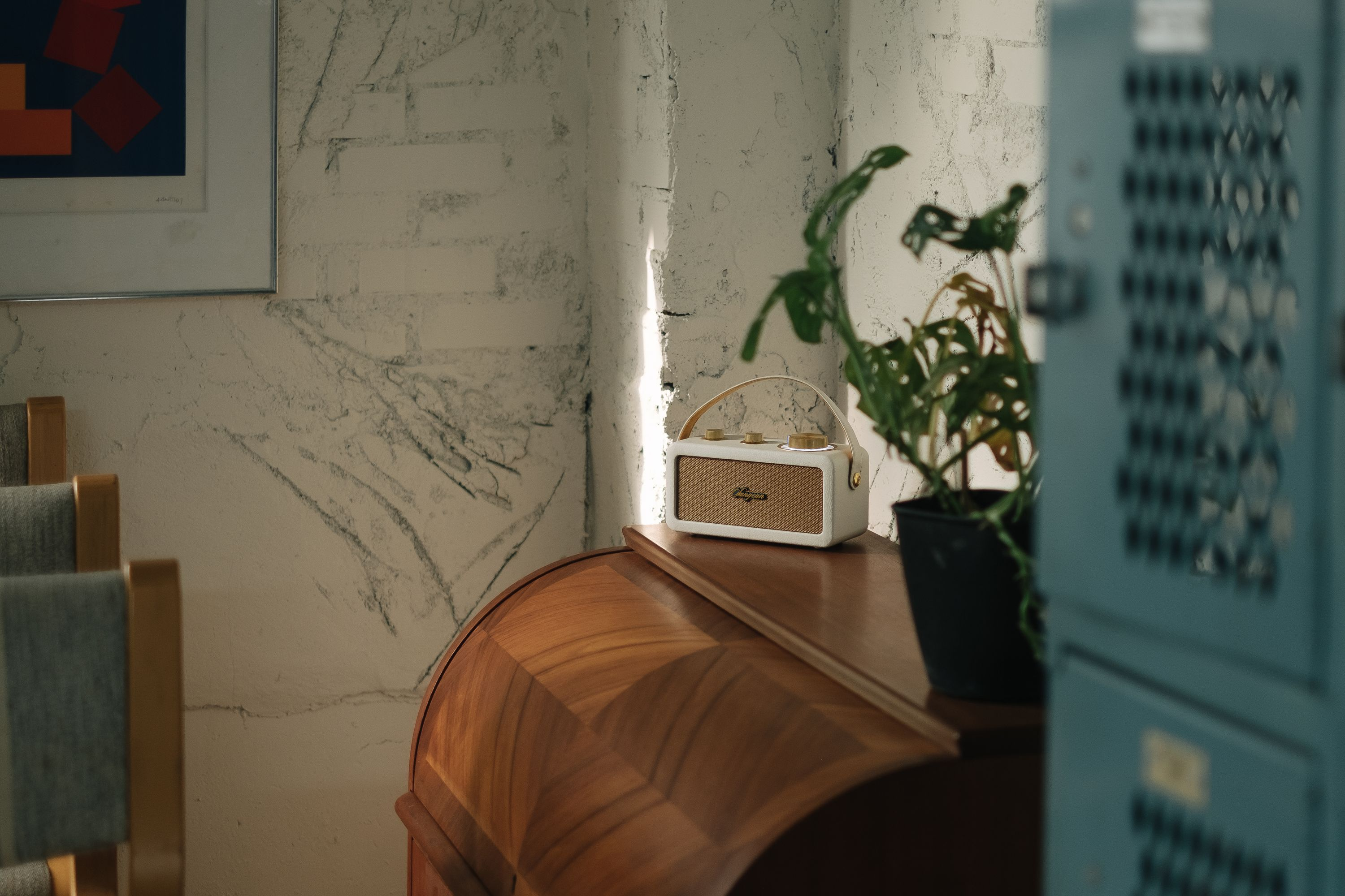 Una radio blanca sobre una superficie de madera, con una planta y taquillas al fondo. Un cuadro cuelga en la pared.