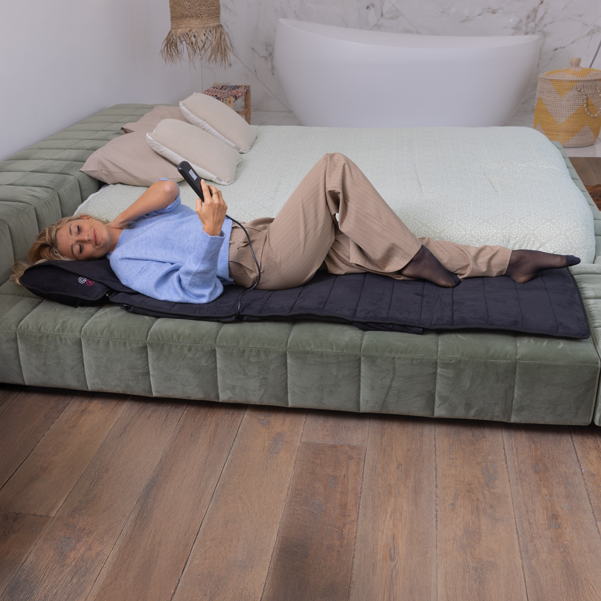 A woman lies on a bed with a massage mat. She holds a device. The room includes a bathtub and a wooden floor.