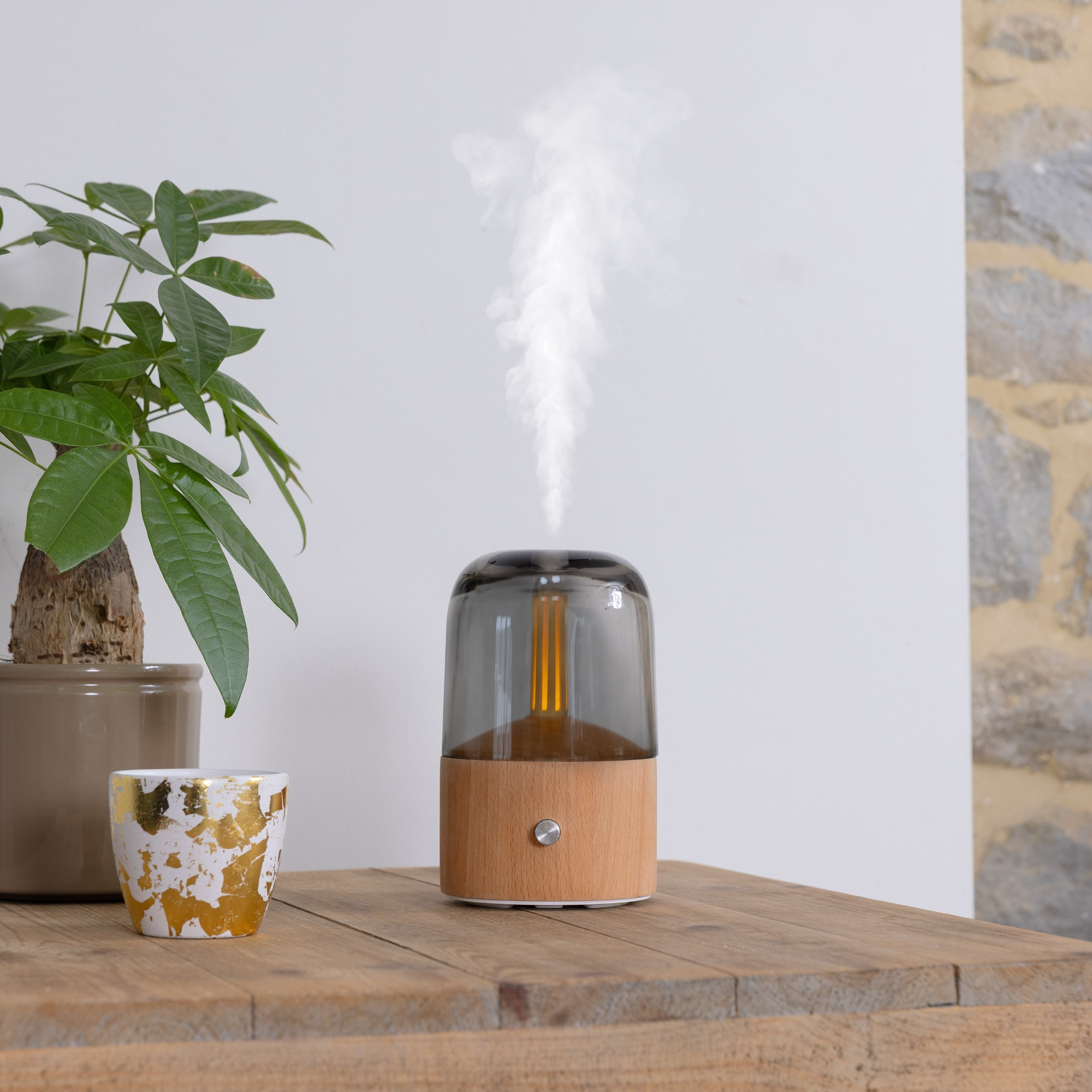 An aroma diffuser on a wooden table with a plant and a small decorated pot. Smoke rises from the diffuser.
