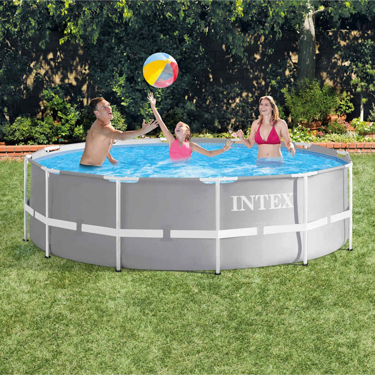 Drei Personen spielen in einem Pool. Ein Mädchen und ein Mann greifen nach einem Ball. Eine Frau im roten Bikini lächelt. Der Pool steht auf grünem Gras.