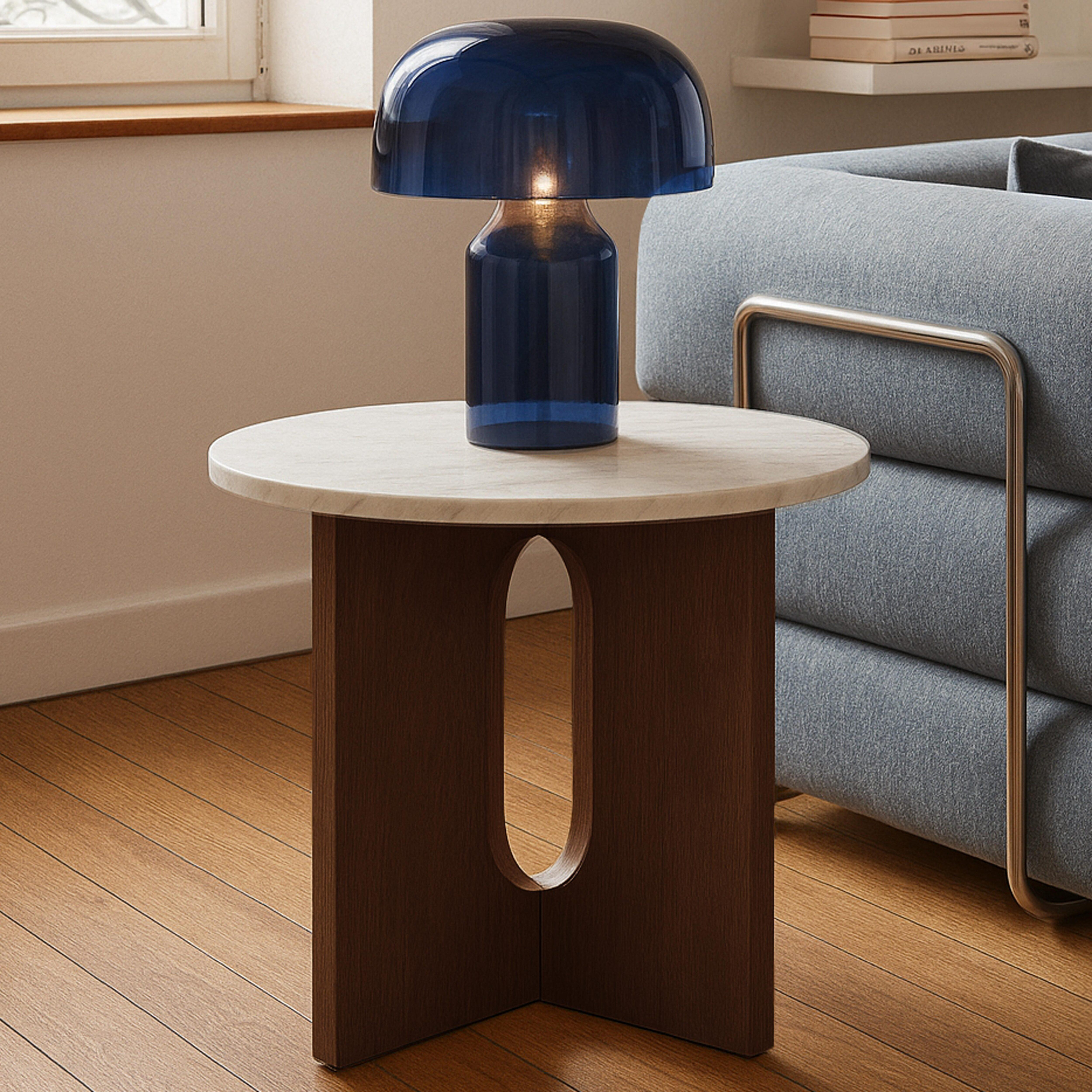 Eine blaue Glaslampe auf einem runden Tisch mit Holzsockel. Graues Sofa und Holzboden im Hintergrund.