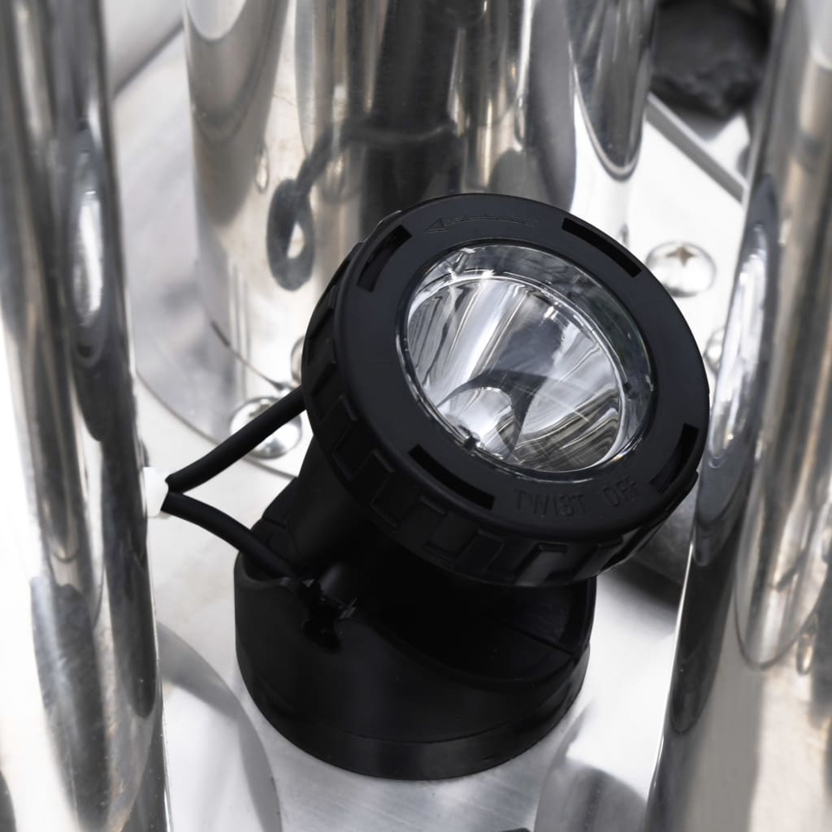 Ein schwarzer Scheinwerfer mit klarer Linse ist auf einer glänzenden Metalloberfläche montiert. Kabel verlaufen von der Leuchte.