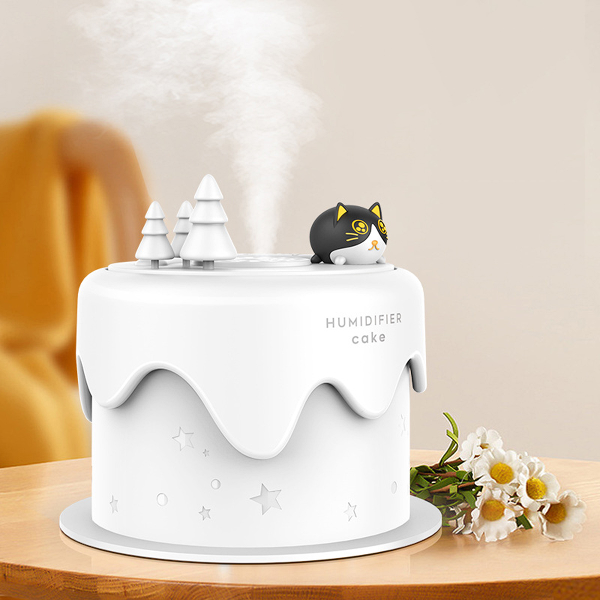 Ein weißer, kuchenförmiger Luftbefeuchter mit einer Katzenfigur, kleinen Bäumen und Dampf. Blumen sind rechts.