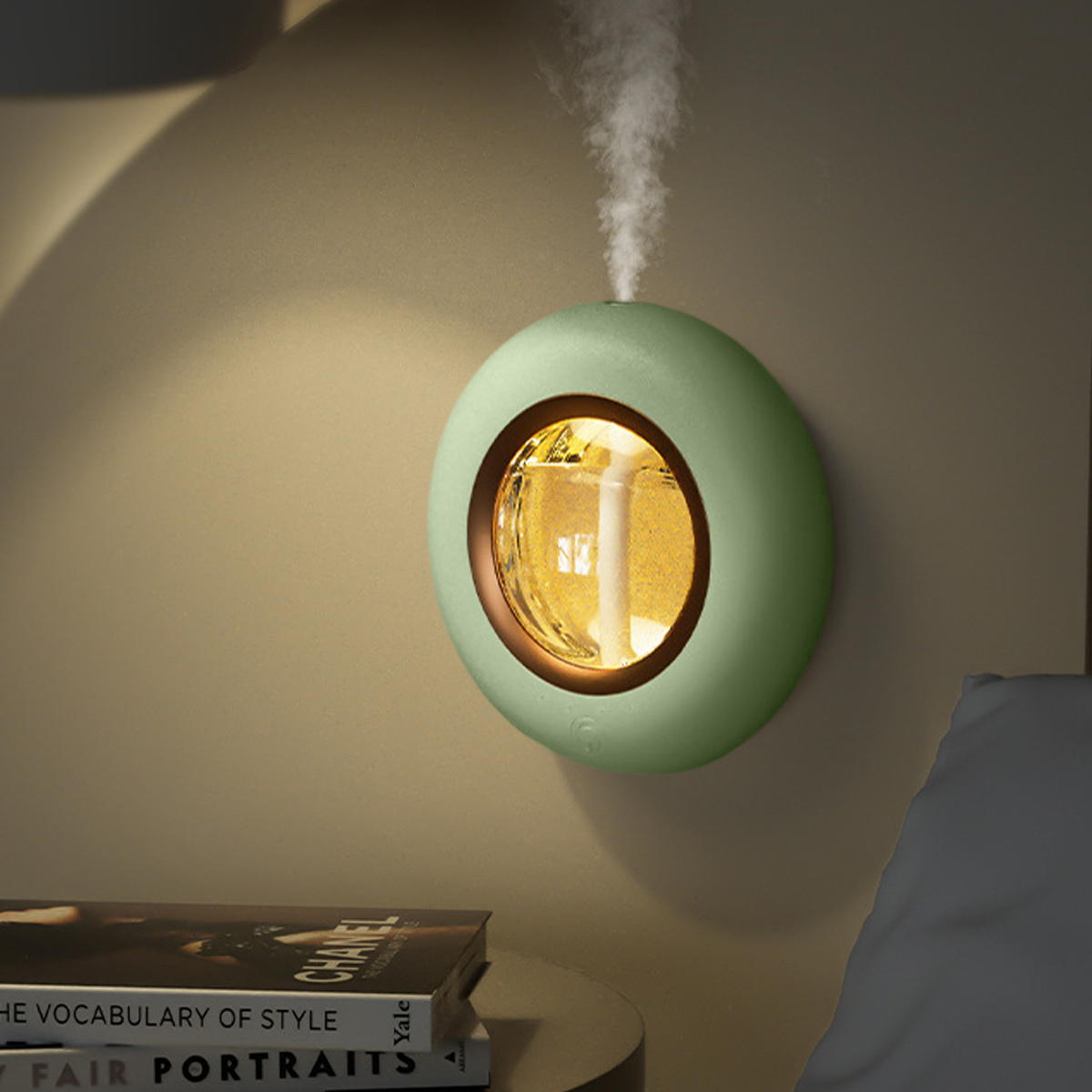 Ein grüner, oval geformter Diffusor für ätherische Öle an einer Wand. Dampf steigt auf. Bücher und ein Tisch sind im Vordergrund. Ein Bett ist im Hintergrund.