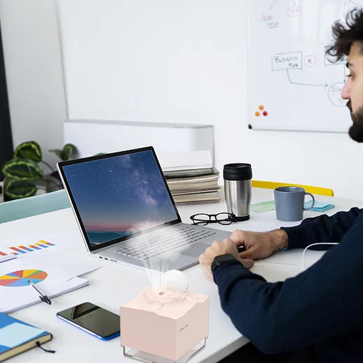 Ein Mann sitzt an einem Schreibtisch. Ein Laptop zeigt einen Sternenhimmel. Ein rosa Luftbefeuchter steht davor. Im Hintergrund befindet sich eine Tafel.