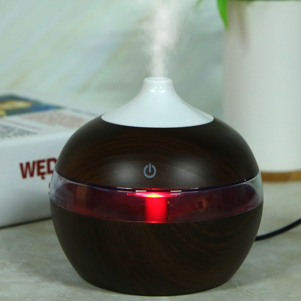 Ein dunkler Holz-Aroma-Diffusor mit weißem Aufsatz, der Dampf abgibt. Er hat ein rotes Licht und einen Power-Knopf. Buch und Pflanze im Hintergrund.