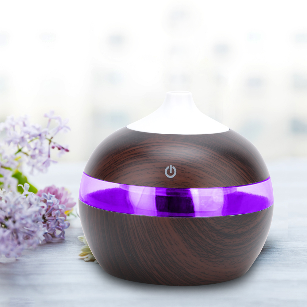 Ein Aroma-Diffusor aus Holz mit einem violetten Lichtband. Links sind Fliederblüten. Der Diffusor hat eine weiße Oberseite und einen Ein-/Ausschalter.