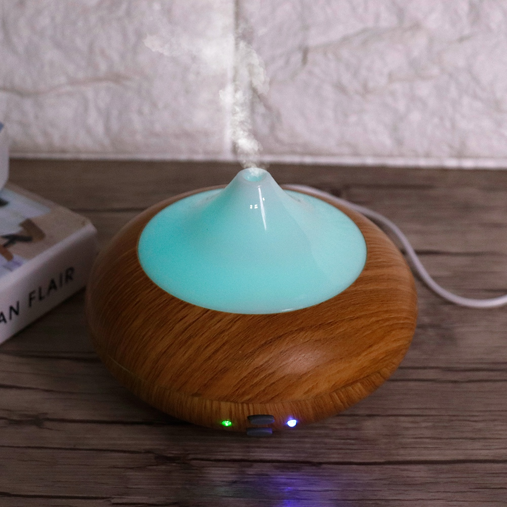 Ein Aroma-Diffusor aus Holz mit tealfarbener Oberseite gibt Dampf ab. Er steht auf Holz, in der Nähe eines Buches. Im Hintergrund eine weiße Backsteinmauer.
