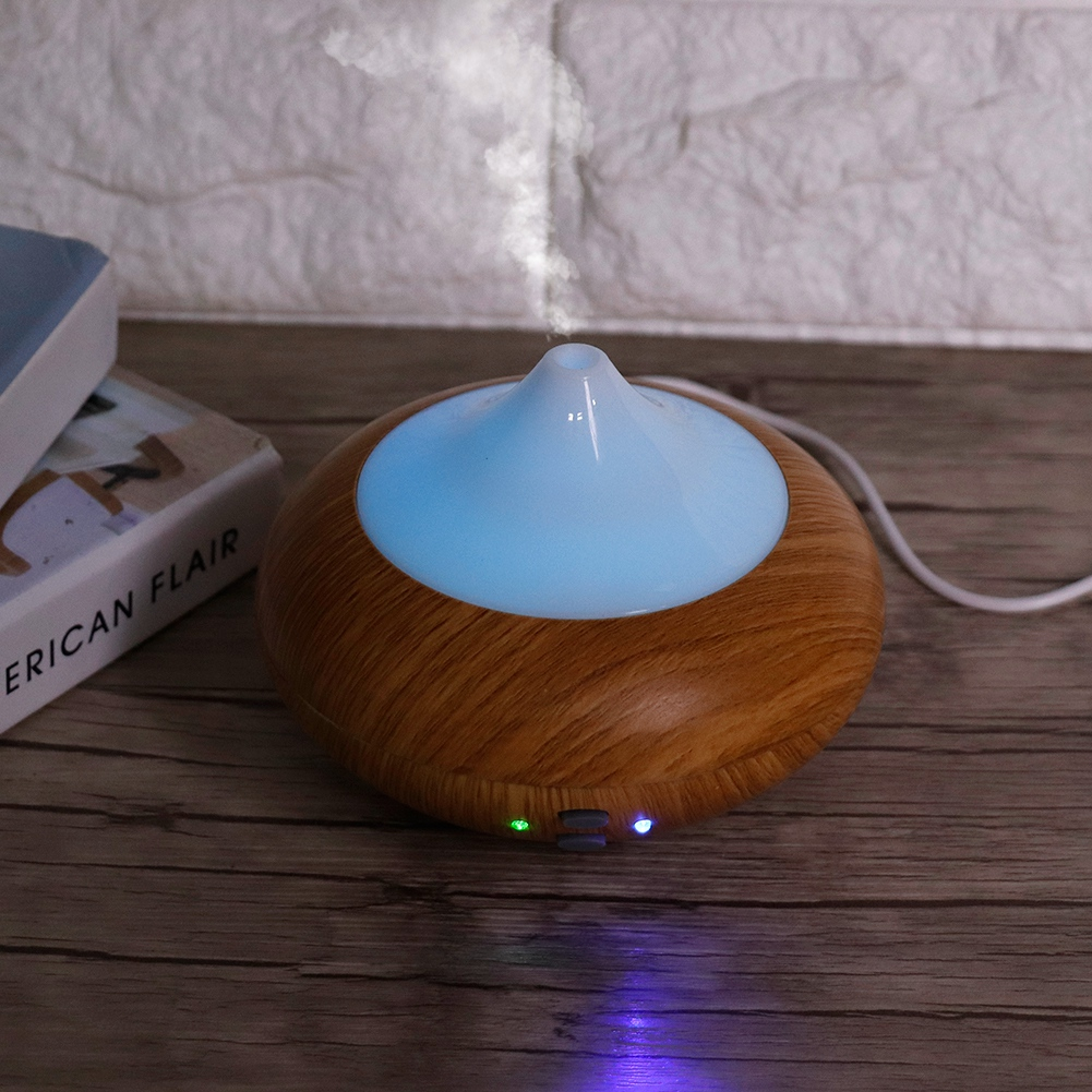 Ein Aromadiffusor steht auf einem Holztisch. Er hat einen Holzmaserungsfuß und eine hellblaue Oberseite, die Dampf abgibt. Bücher sind im Hintergrund.